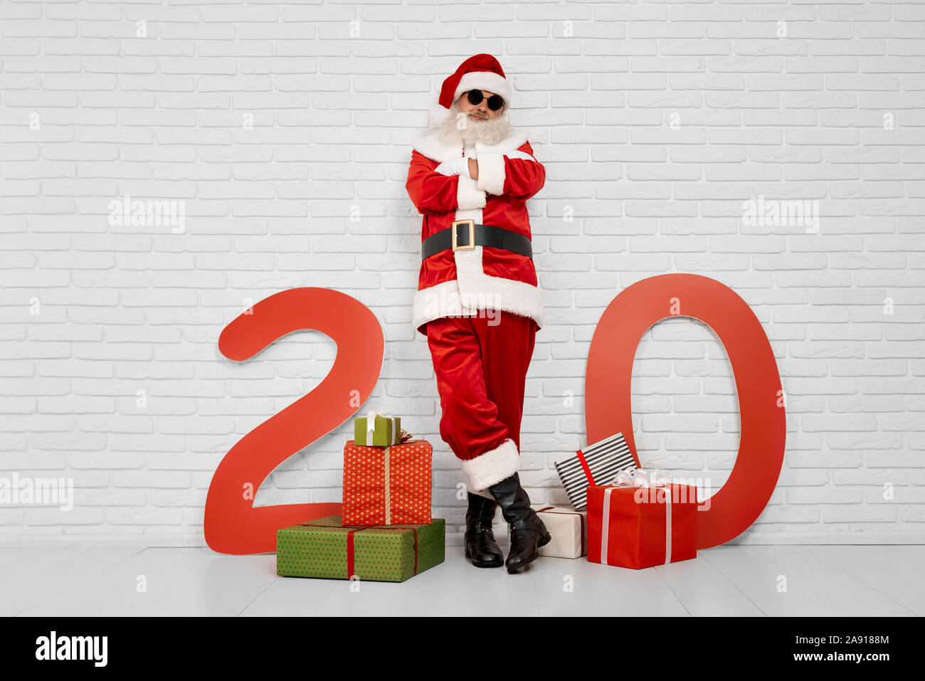 Cool Santa Claus in schwarz Sonnenbrille lean gegen weiße Wand zwischen wunderschöne Geschenkboxen und Papier rot gefärbt Anzahl von 20. Gerne älterer Mann ist bereit, neue Jahr Party zu feiern. Stockfoto