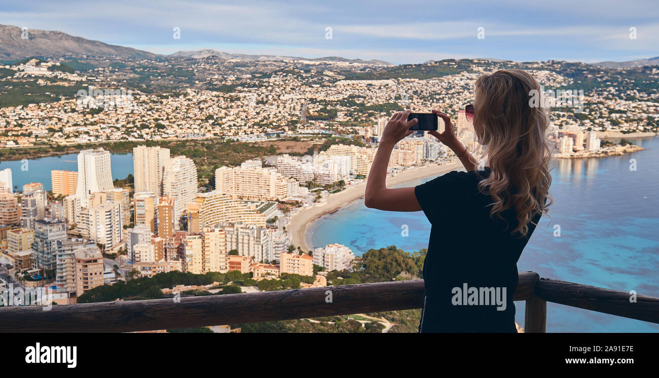 Junge blonde Frau steht auf eingezäunten Bereich kletterte auf Penon de Ifach Symbol von Calpe spanischen touristischen Stadt, hält Smartphone dauert Bild der Landschaft Stockfoto