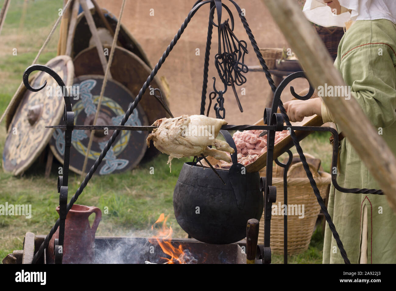 Frau gekleidet in mittelalterlichen Kostüm Vorbereiten der Nahrung zu einem Re-enactment Camp mit einem Spieß gebratenes Huhn und Kessel Stockfoto