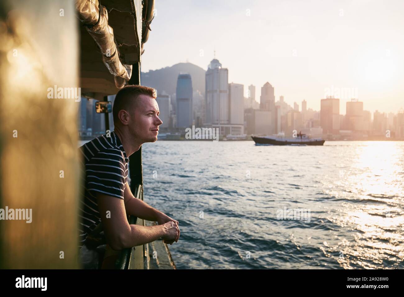 Junger Mann (Reisenden) von Boot gegen moderne Stadt am wunderschönen Sonnenuntergang. Stadtbild mit Wolkenkratzern in Hongkong. Stockfoto