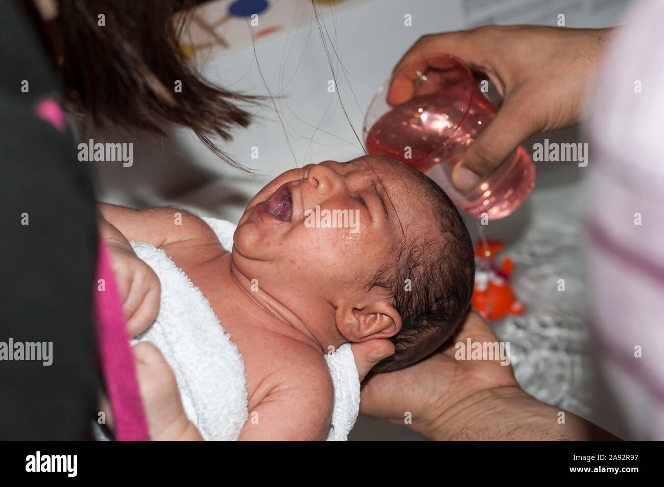 Glas mit Wasser über den Kopf des Weinens zwei Wochen alten Säugling seine Haare zu waschen gegossen wird. Baby gehalten von der Mutter und von der Hand des Vaters holding Glas gewaschen. Stockfoto