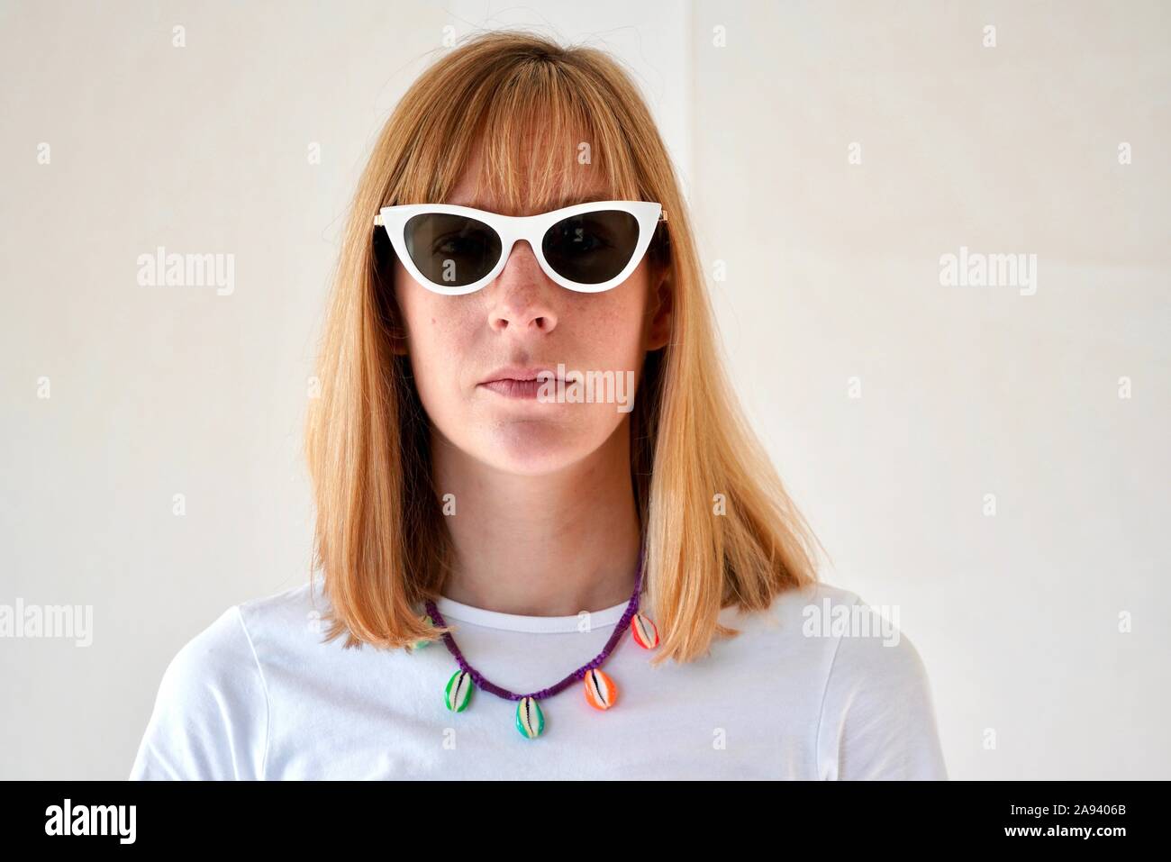 Portrait junge blonde Frau mit neutralen Ausdruck tragen weiße Katze - Auge Sonnenbrille und ein Puka Shell Halskette Stockfoto