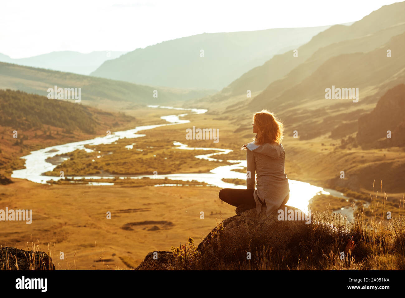 Schlanke Mädchen sitzt auf Big Rock und genießt die Aussicht auf den Sonnenuntergang Berge und den Fluss Stockfoto