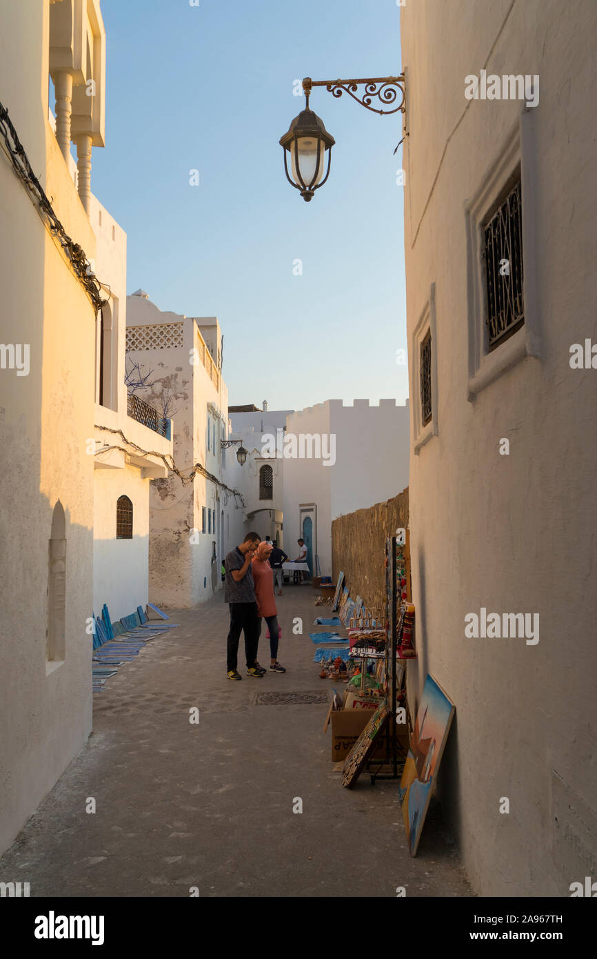Asilah, Morocco-September 10, 2019: Alte Straße in der Medina, der Altstadt von Asilah mit Touristen Engen an Gemälde, die für Verkauf suchen, Marokko Stockfoto