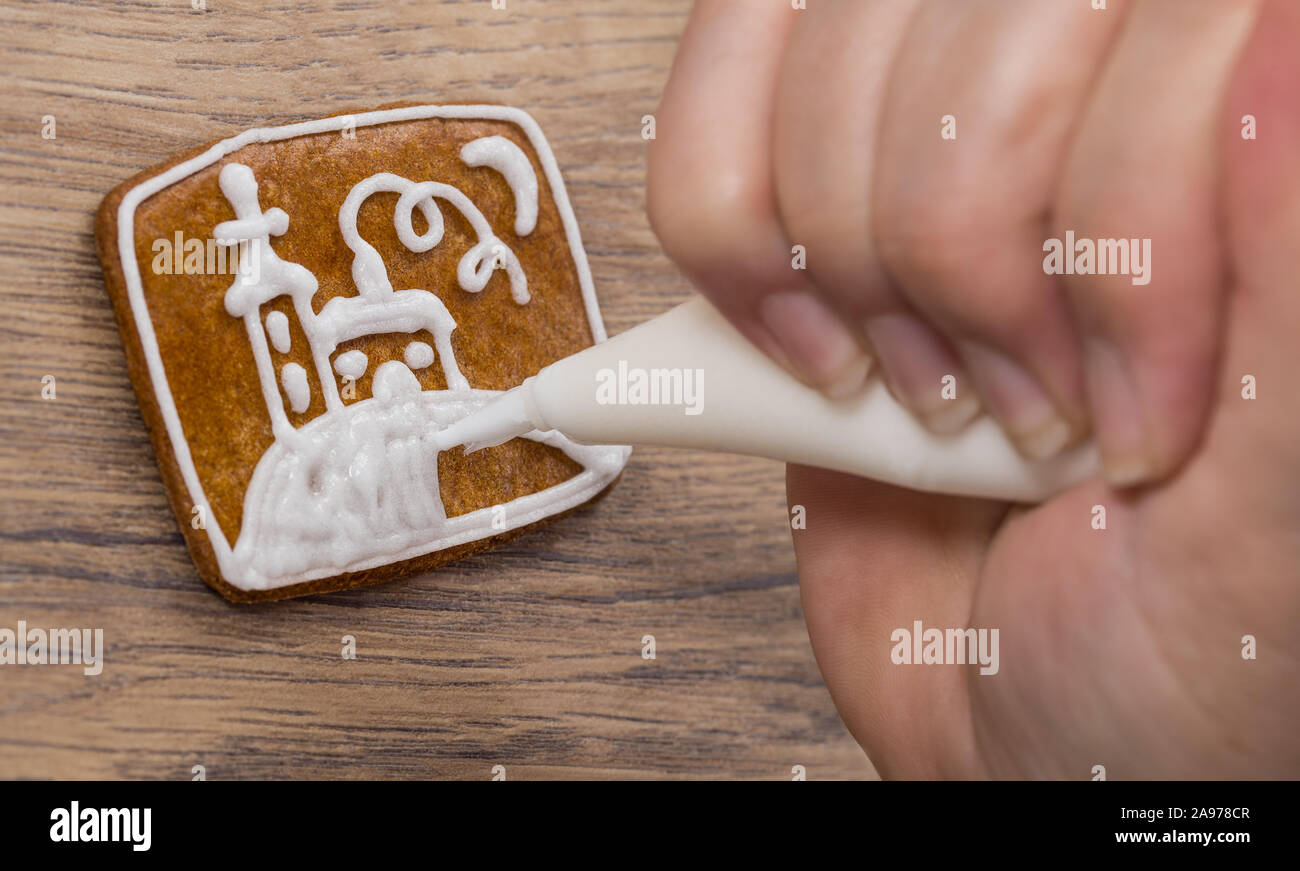 Menschliche Hand dekorieren Süße Weihnachten Lebkuchen mit religiösen Thema. Gebäckbeutel bei weiblichen Finger malen eine weisse Kirche zu Brown cookie gepresst. Stockfoto