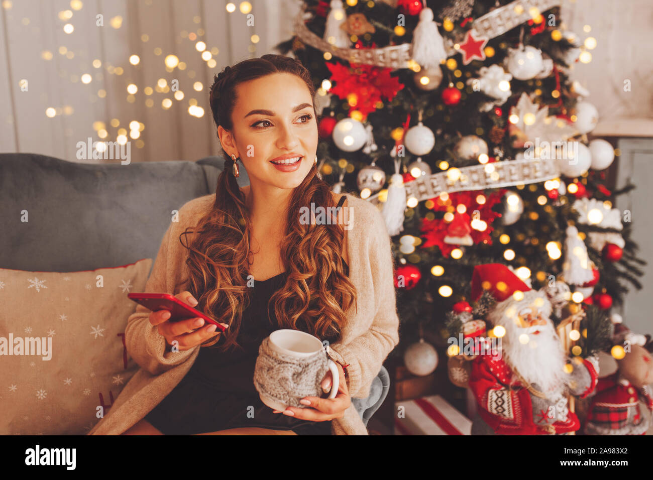 Gerne Frau sitzt auf dem Sofa und Blick auf Fenster und Handy halten. Weihnachtsbaum mit Dekoration ist neben ihr. Ferien- und Technologiekonzepte. Stockfoto