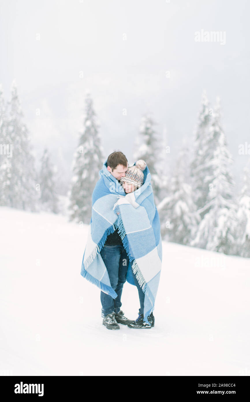 Jungen gutaussehenden Mann umarmt seine hübsche junge Mädchen und stopft sie mit der Wolle chekered blaue Decke. Winterwanderung im verschneiten Wald. Liebe im Winter Stockfoto