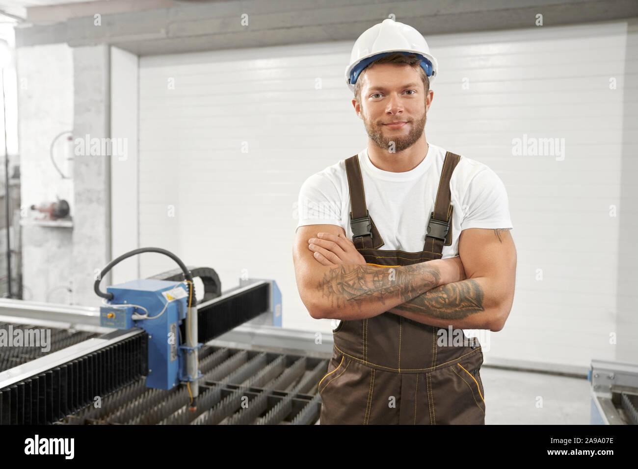 Vorderansicht des gutaussehenden Mann in Uniform und Helm stehen mit verschränkten Armen, Kamera und lächelnd. Männliche Mechaniker arbeiten mit CNC-auf Metall Werk. Konzept der Maschinen. Stockfoto