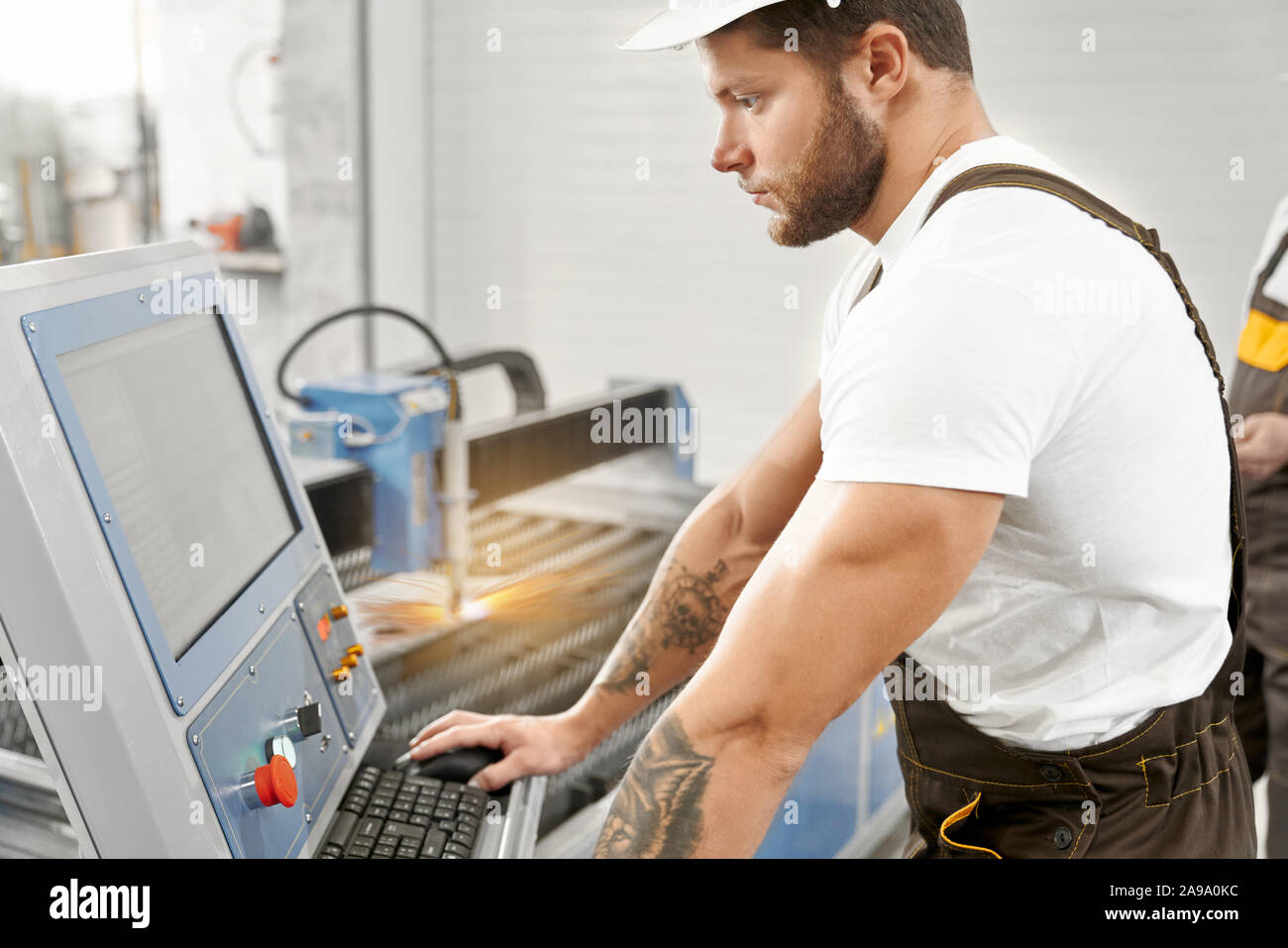 Ansicht von der Seite des jungen Mann in Uniform und Helm arbeiten mit professioneller Ausstattung und Computer auf Fabrik, männliche Schweißer controlling Prozess der Plasmaschneiden. Konzept der Maschinen. Stockfoto