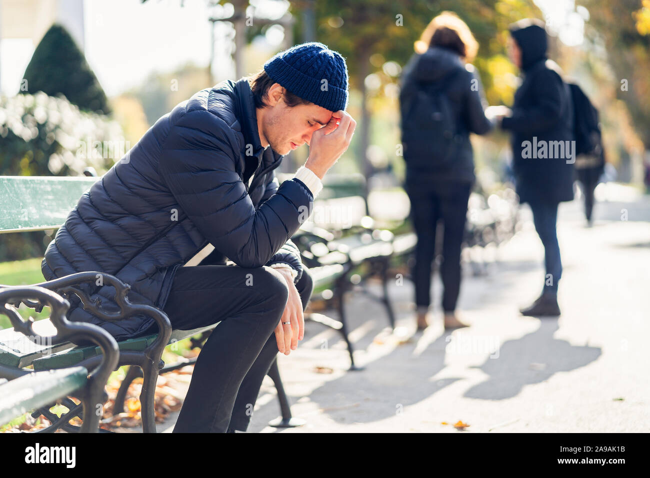 Junge besorgt, der Mensch ist in seinem Gesicht. Er sitzt auf einer Bank während der sonnigen Herbsttag. Der Menschen silhuettes im Hintergrund sichtbar. Stockfoto