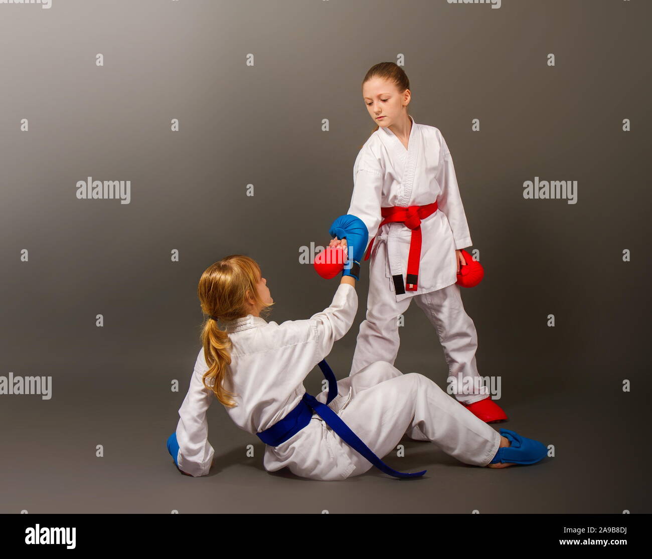 Zwei kleine Karate Mädchen in einem Kimono und voller Sport Schutzausrüstung von roten und blauen Farben helfen, ein Gegner, der in der Schlacht zu klettern gefallen ist. Stockfoto