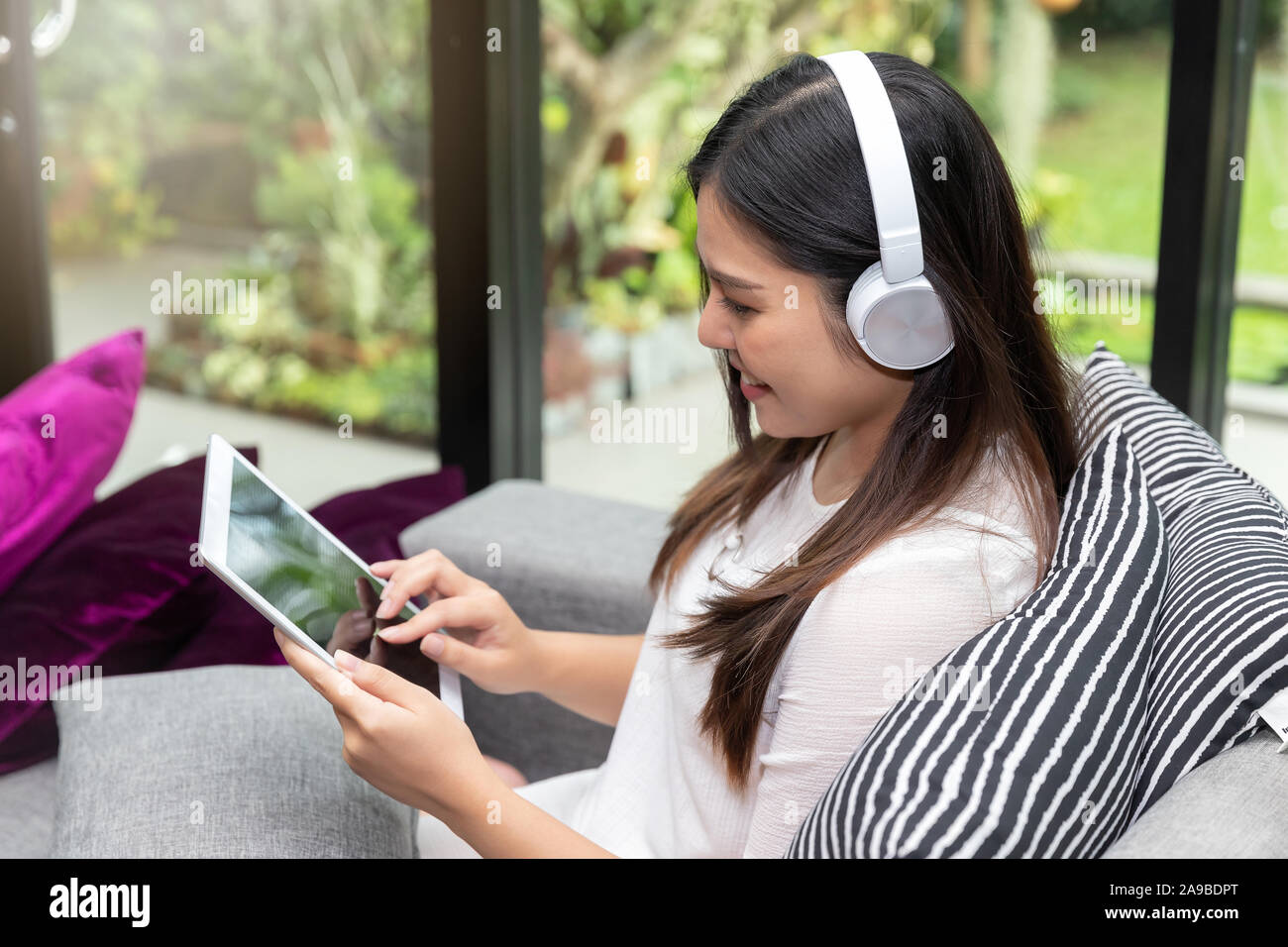 Ziemlich lächelnden jungen Frau tragen Kopfhörer sitzen auf dem Sofa und die Verwendung auf dem Tablet. Stockfoto