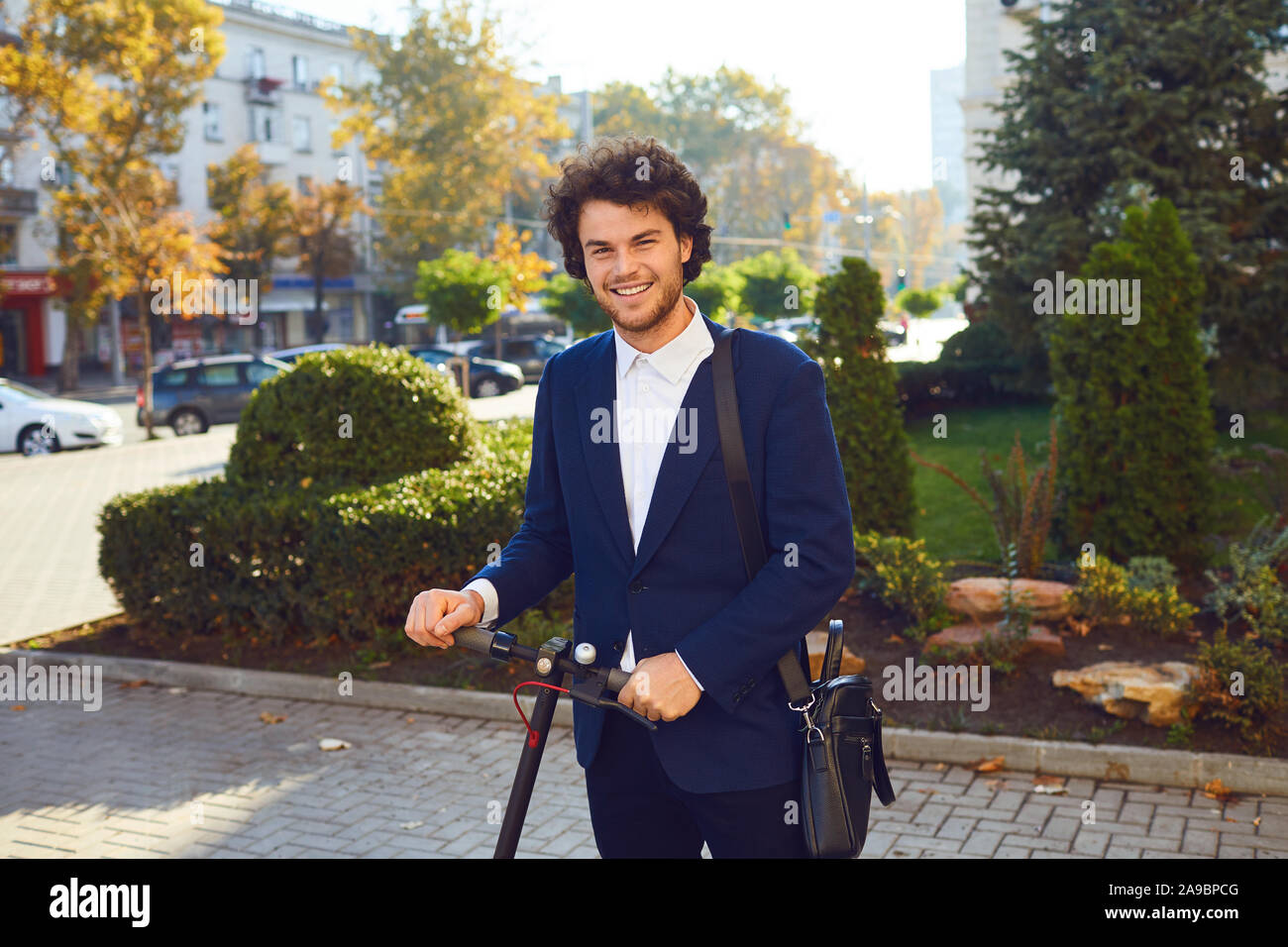 Ein lächelndes curly Kerl steht mit einem elektrischen scooter auf einer Straße der Stadt. Stockfoto