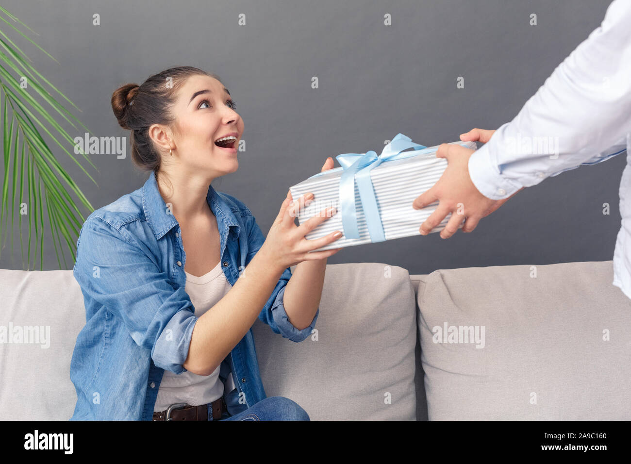 Urlaub Konzept. Junges Paar Mann, geben, Geschenk, Frau sitzt auf einem Sofa lächelnd überrascht Studio auf Grau schließen isoliert - oben Stockfoto
