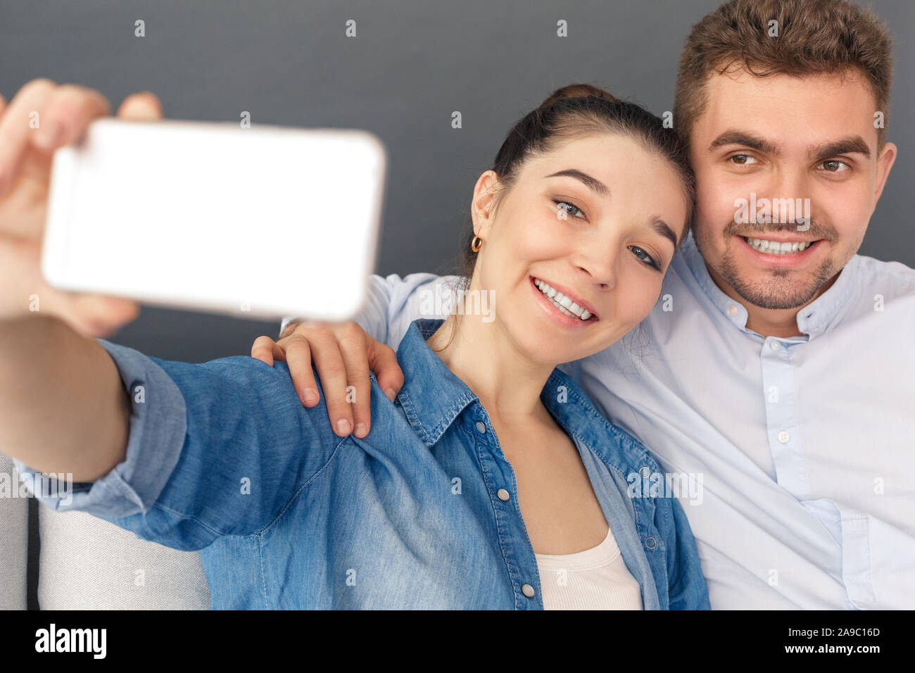 Relationnship Konzept. Junges Paar umarmen Sitzen auf einem Sofa studio isoliert auf Grau unter selfie auf dem Smartphone lächelt fröhlich in die Kamera close-up Stockfoto