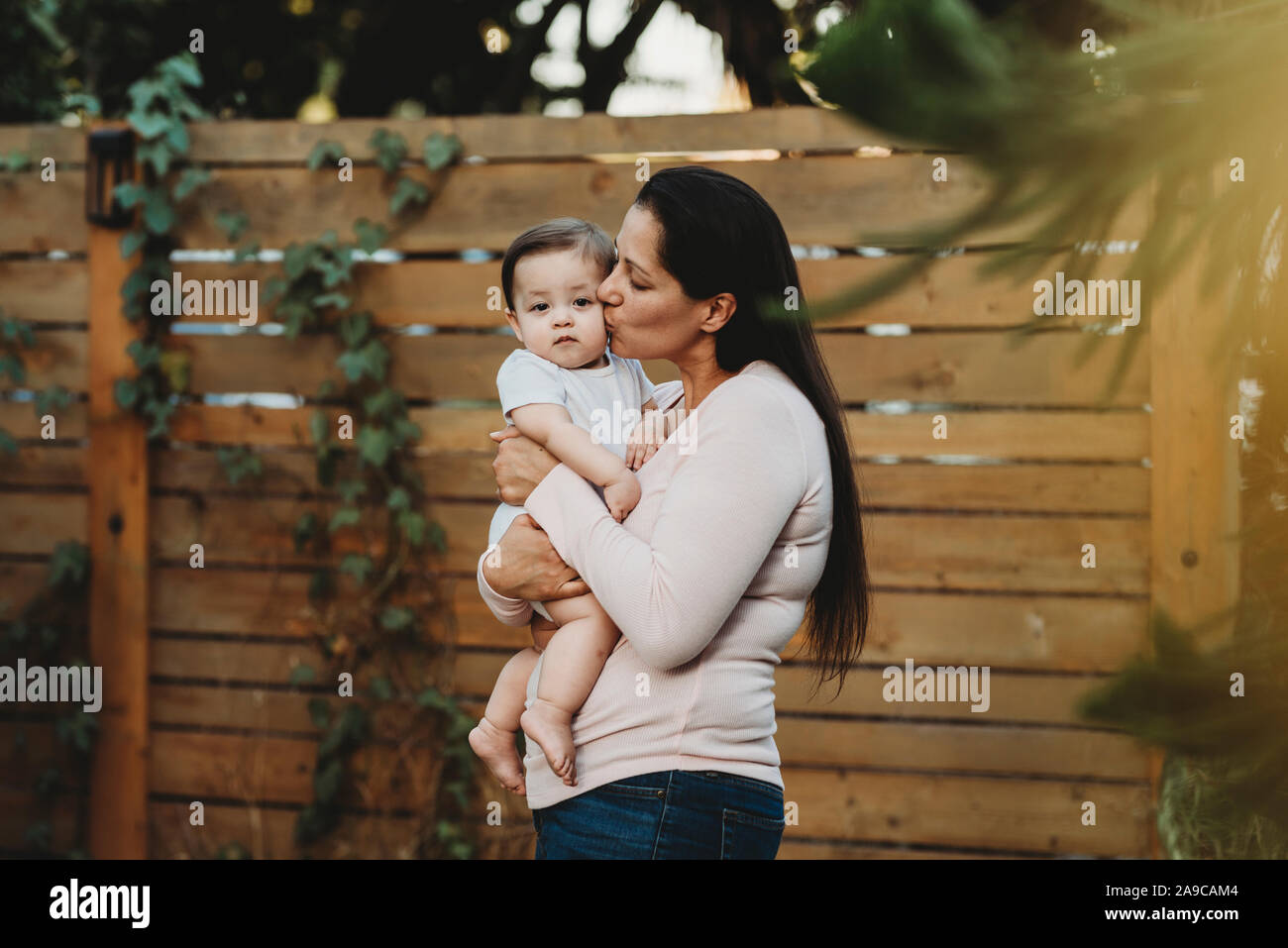 Mitte-30 Mama halten und küssen Baby Mädchen im Hinterhof in der Nähe von Zaun Stockfoto