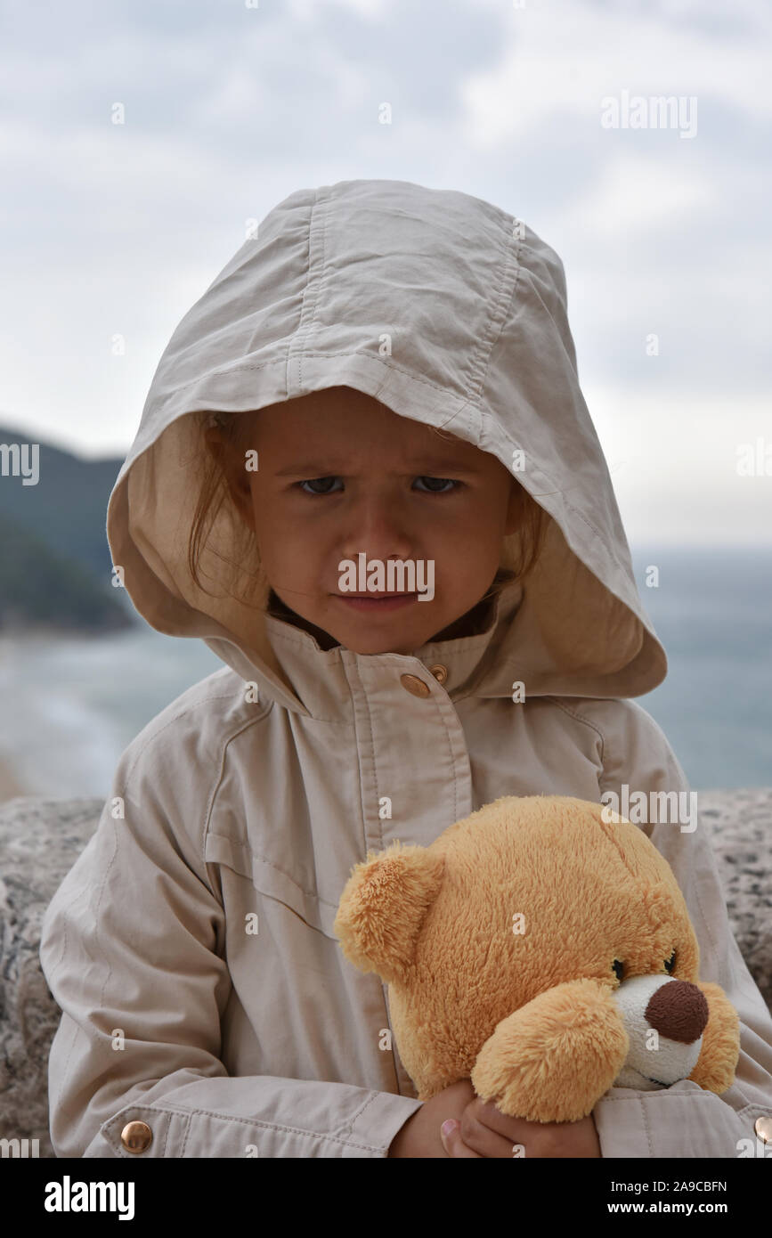 Kleines Mädchen mit Teddybär trägt einen Regenmantel Jacke mit Kapuze gegen einen stürmischen Himmel. Kindheit Konzepte. Ökologische Konzepte. Klima Konzepte. Stockfoto