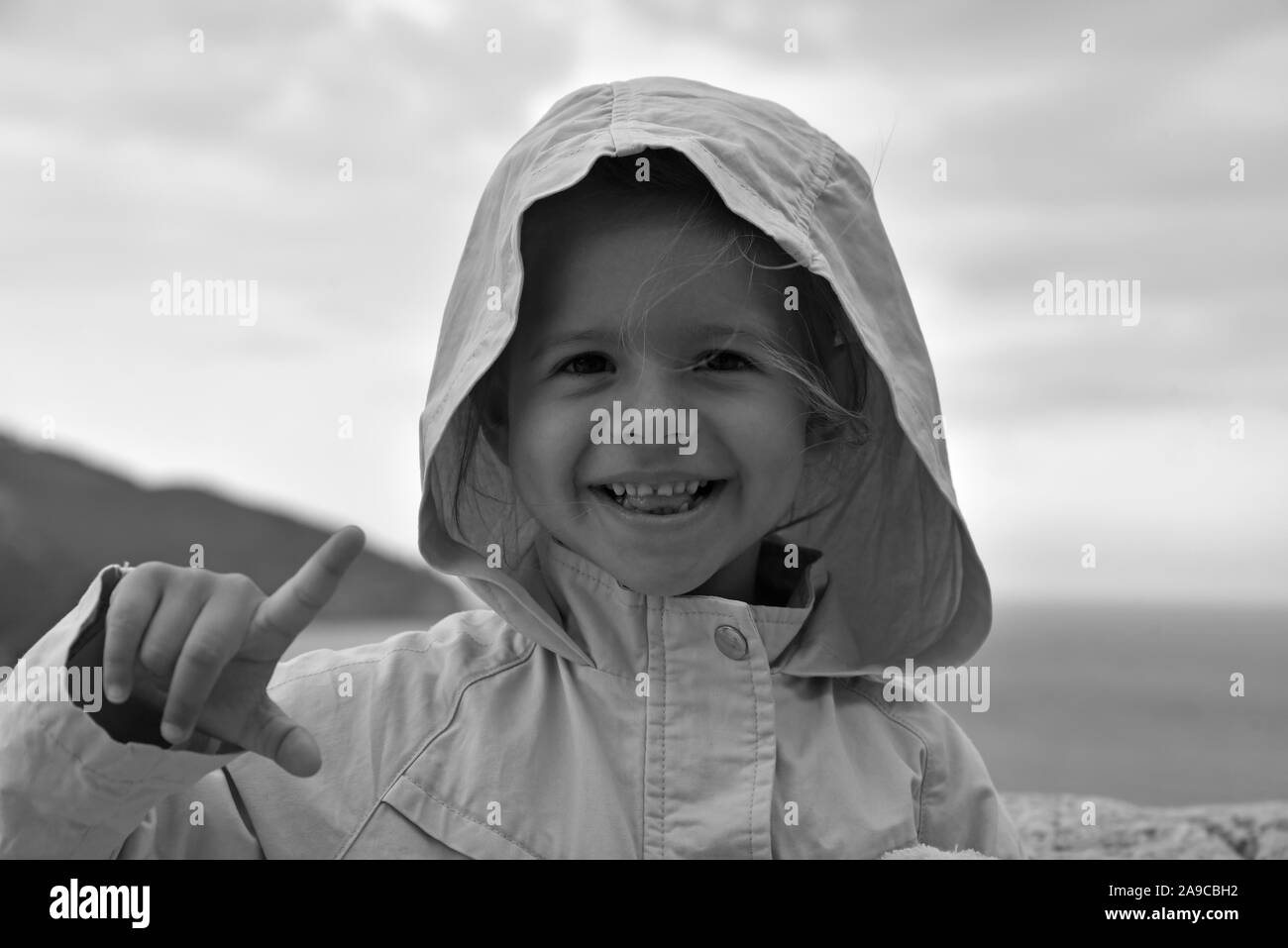 Ein wenig lächelnde Mädchen trägt ein regenmantel Jacke mit Kapuze gegen die stürmischen Himmel. Sie hebt den Zeigefinger. Kindheit Konzepte. Umwelt con Stockfoto
