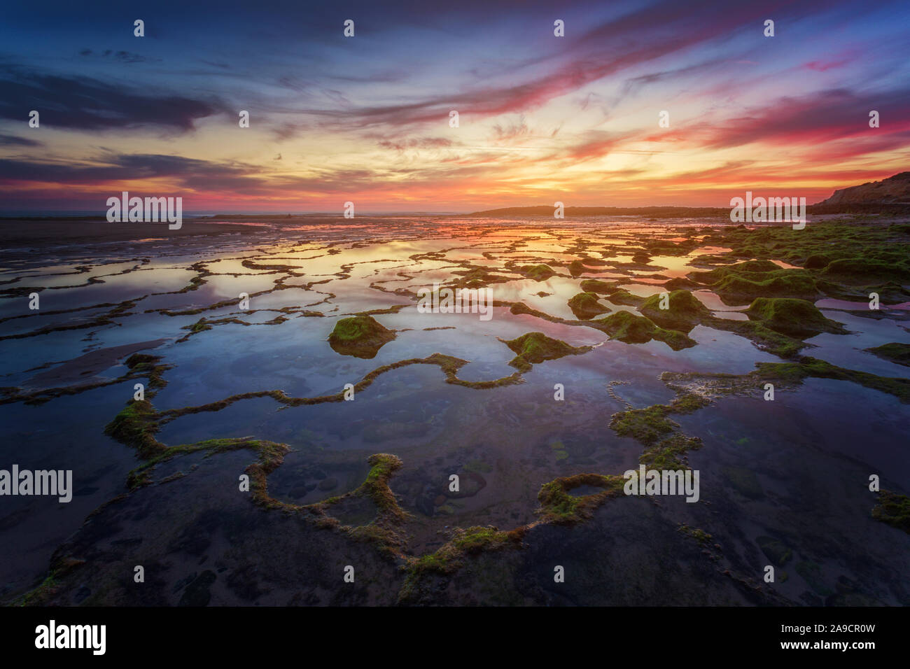 Sonnenuntergang am Strand Praia do Farol, in der Region Alentejo, Portugal. Erste Ebene der Net Shape Felsformation mit Moos und voller Wasser. Meer spiegelt den Himmel Stockfoto