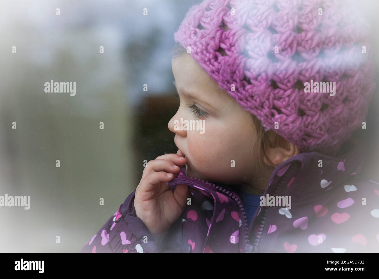 Mädchen mit rosa Mütze und Jacke stehen am Fenster suchen, den Finger auf den Mund, Kopf hoch Stockfoto