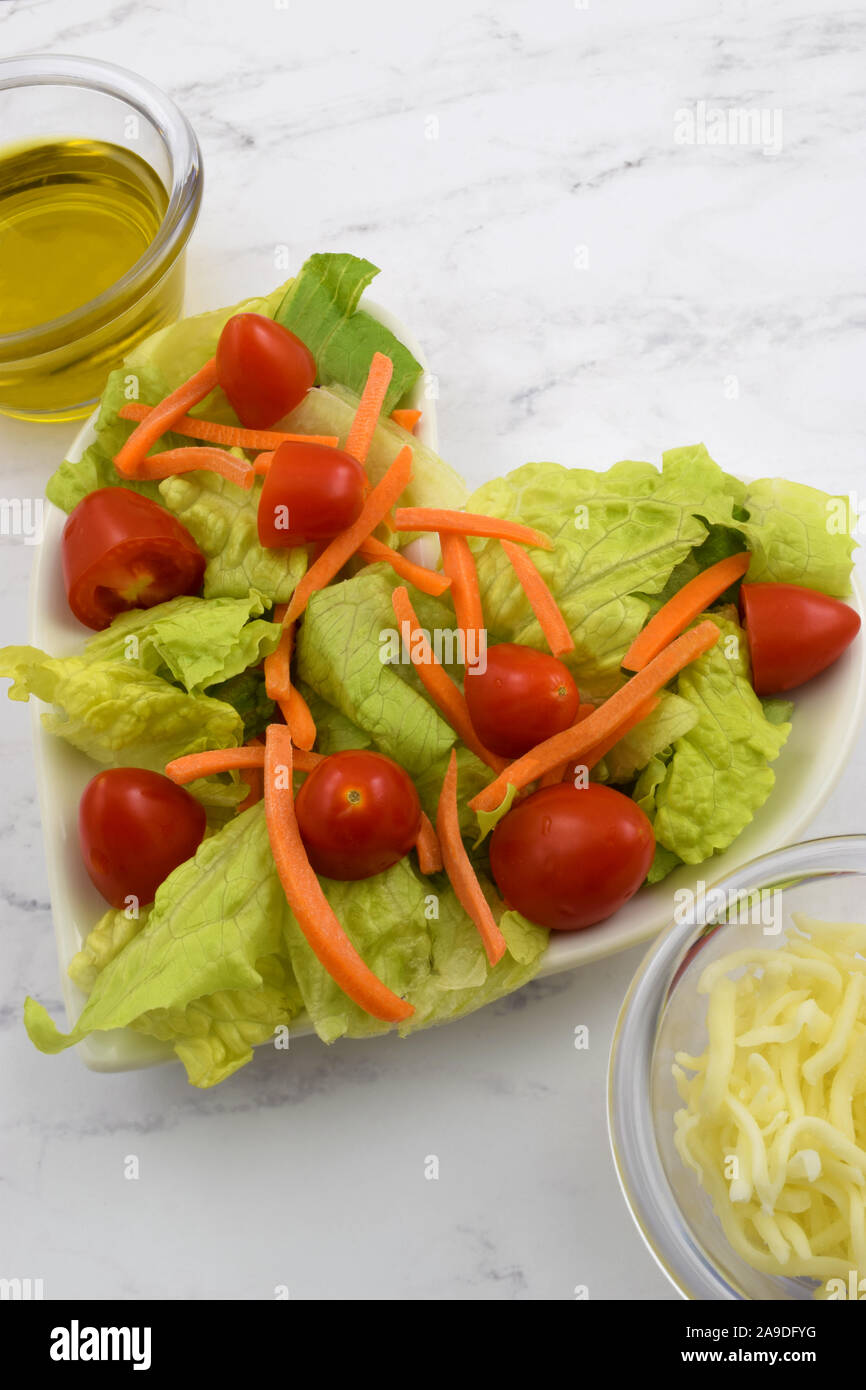 Frische Herz gesunden Salat auf einem herzförmige Platte mit Römersalat, Karotten, Tomaten, Olivenöl und geriebenem Käse. Mit Liebe gemacht. Stockfoto