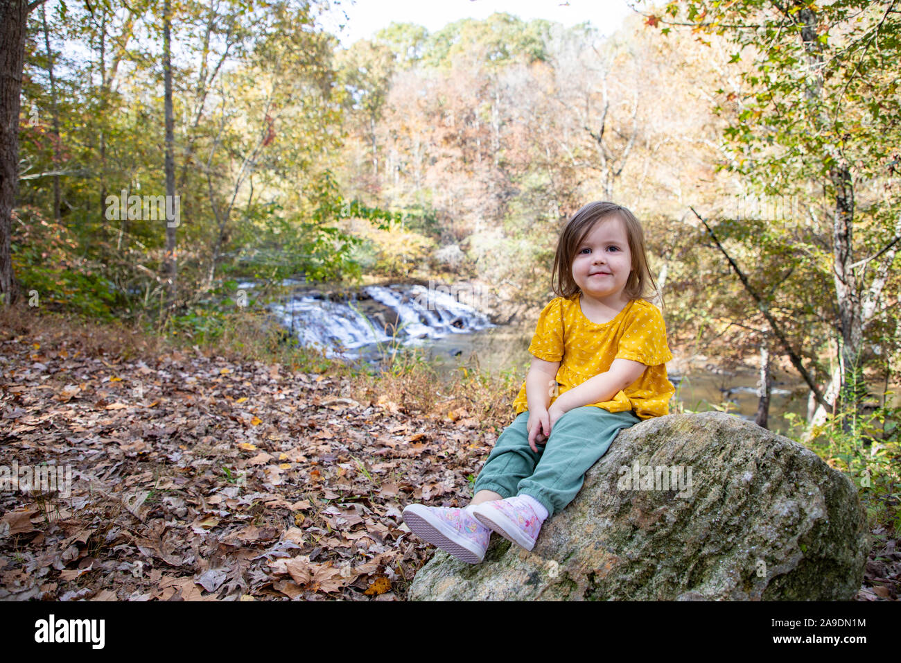 Kleinkind auf Felsen sitzend mit Wasserfall im Hintergrund Stockfoto
