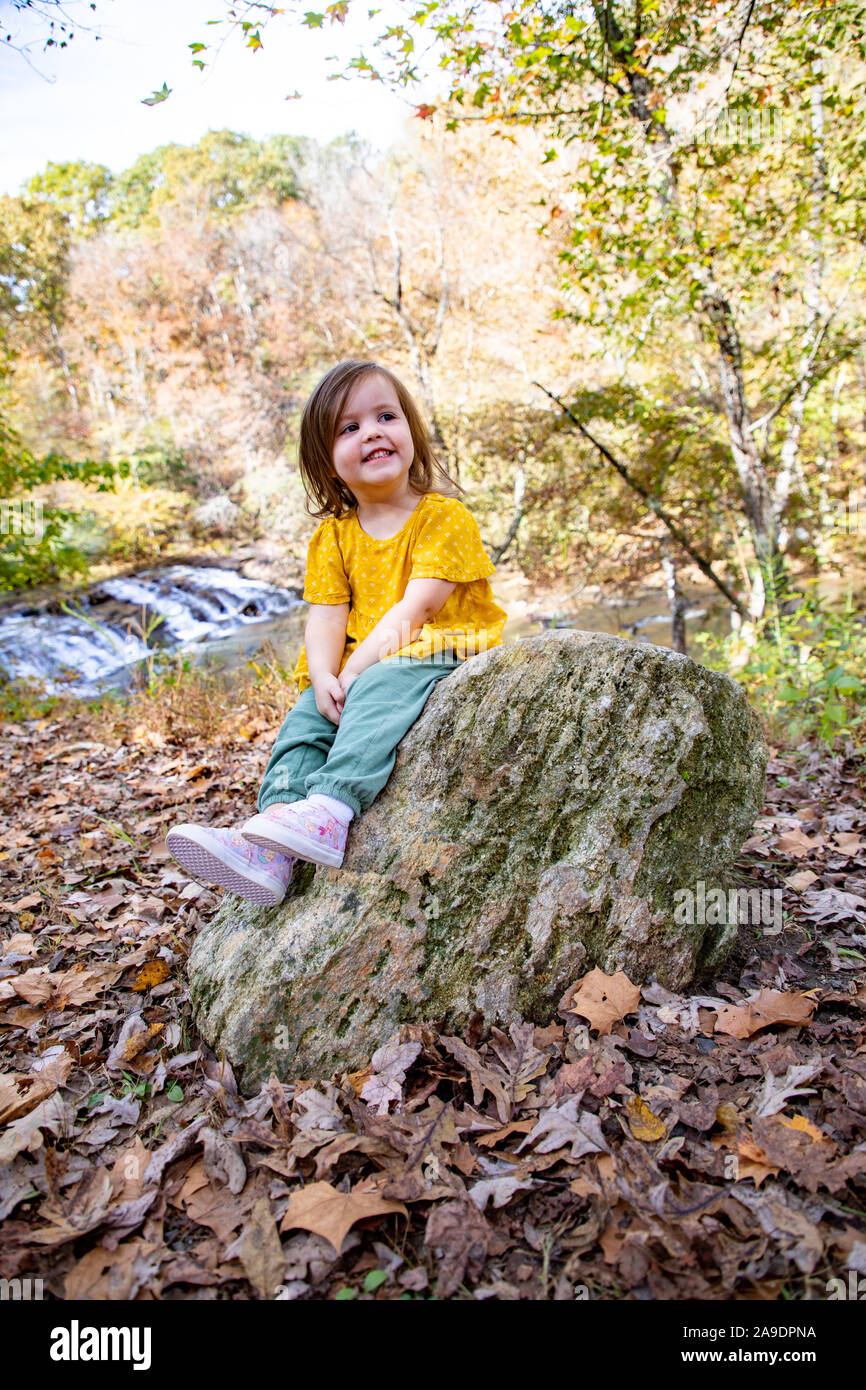 Kleinkind in gelbem Hemd auf Felsen in der Nähe des Wasserfalls - lächelnd Stockfoto