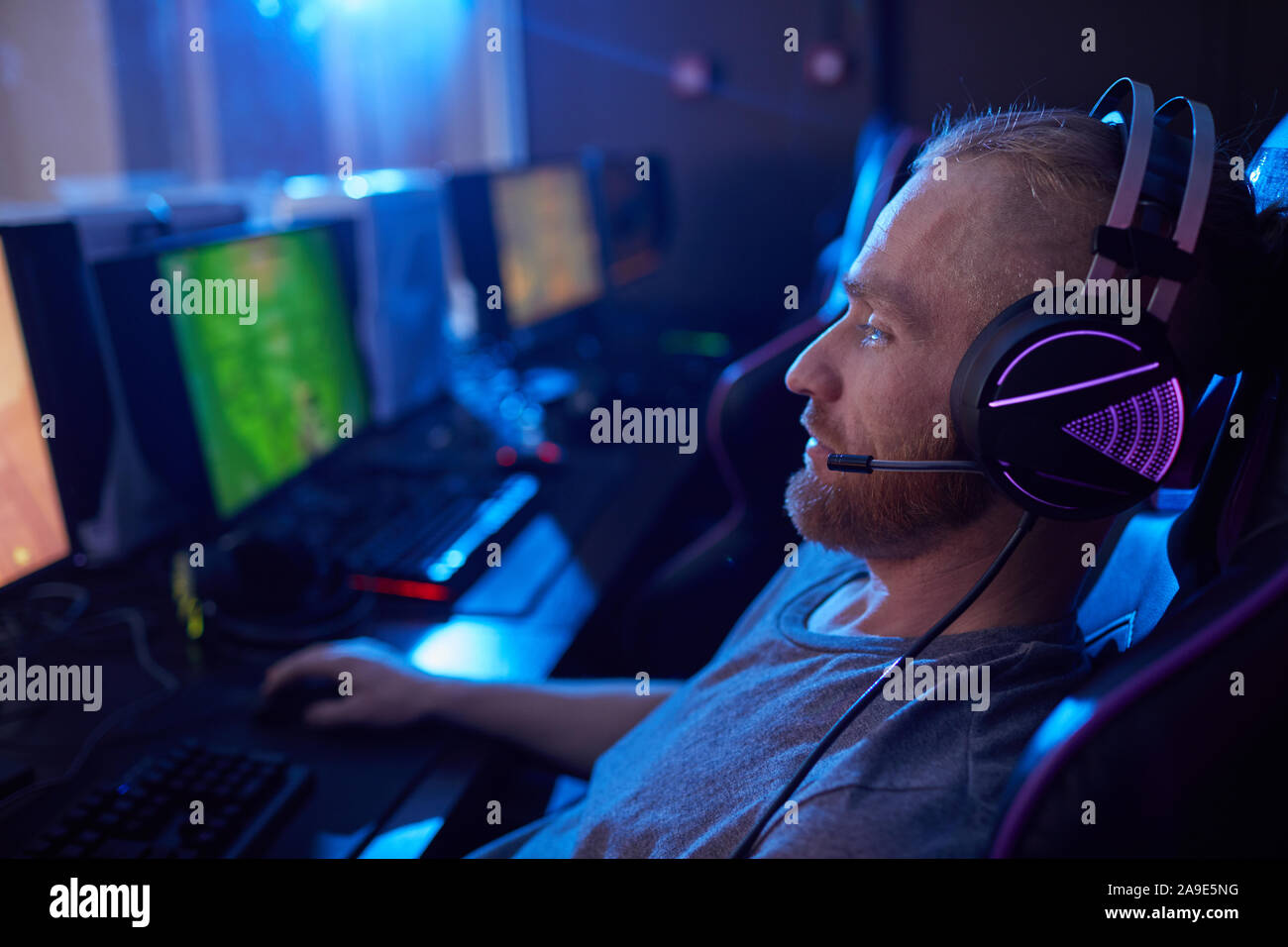 Seitenansicht des jungen Mannes in Gaming Kopfhörer mit Hintergrundbeleuchtung sitzen und spielen im Computer Spiel in dunklen Computer klasse Stockfoto
