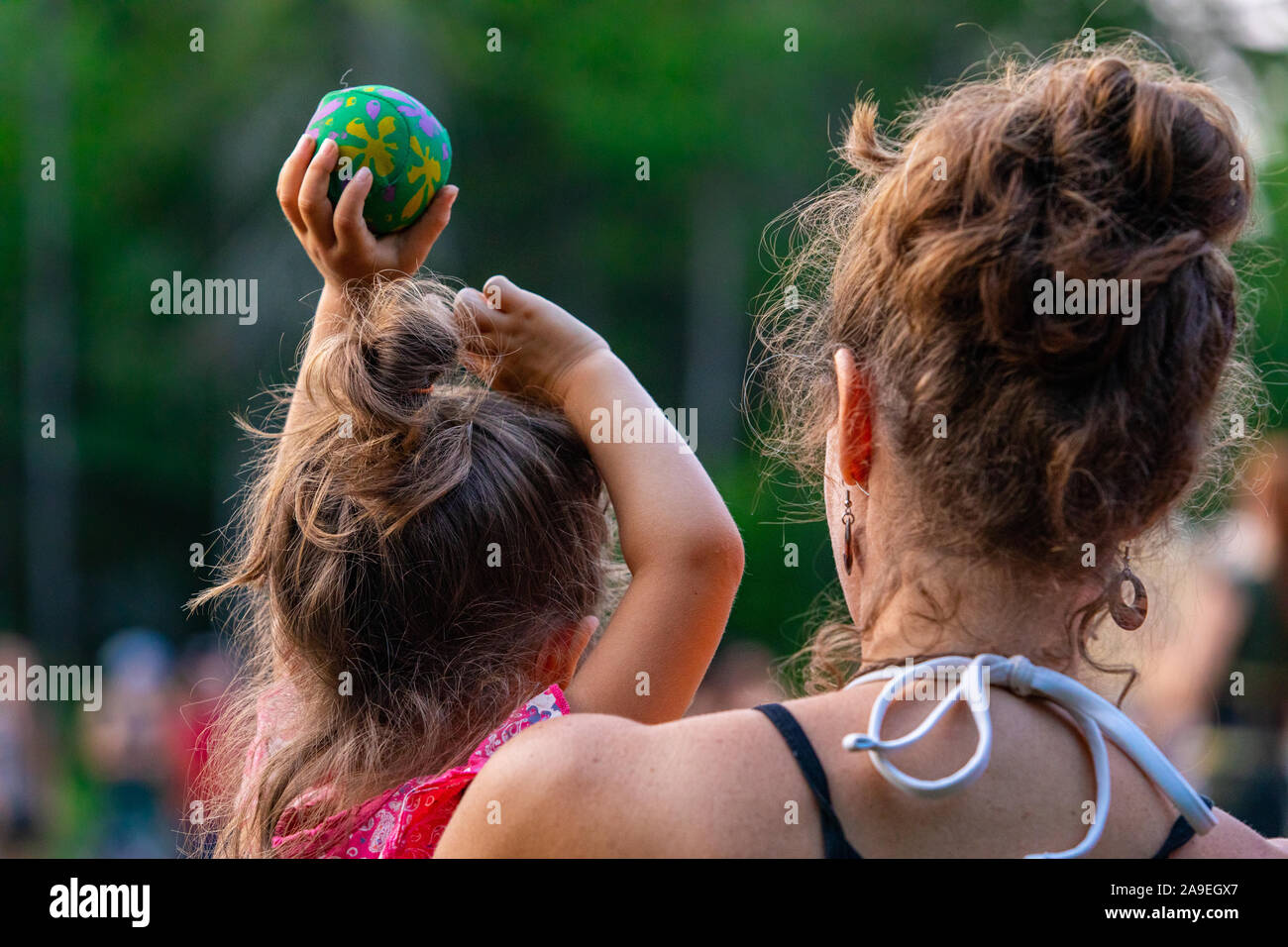 Selektive konzentrieren und auf die junge Mutter hält ihre Tochter in die Arme von hinten gesehen, mit einer Gruppe von Menschen, die im Hintergrund zu sehen sind Stockfoto