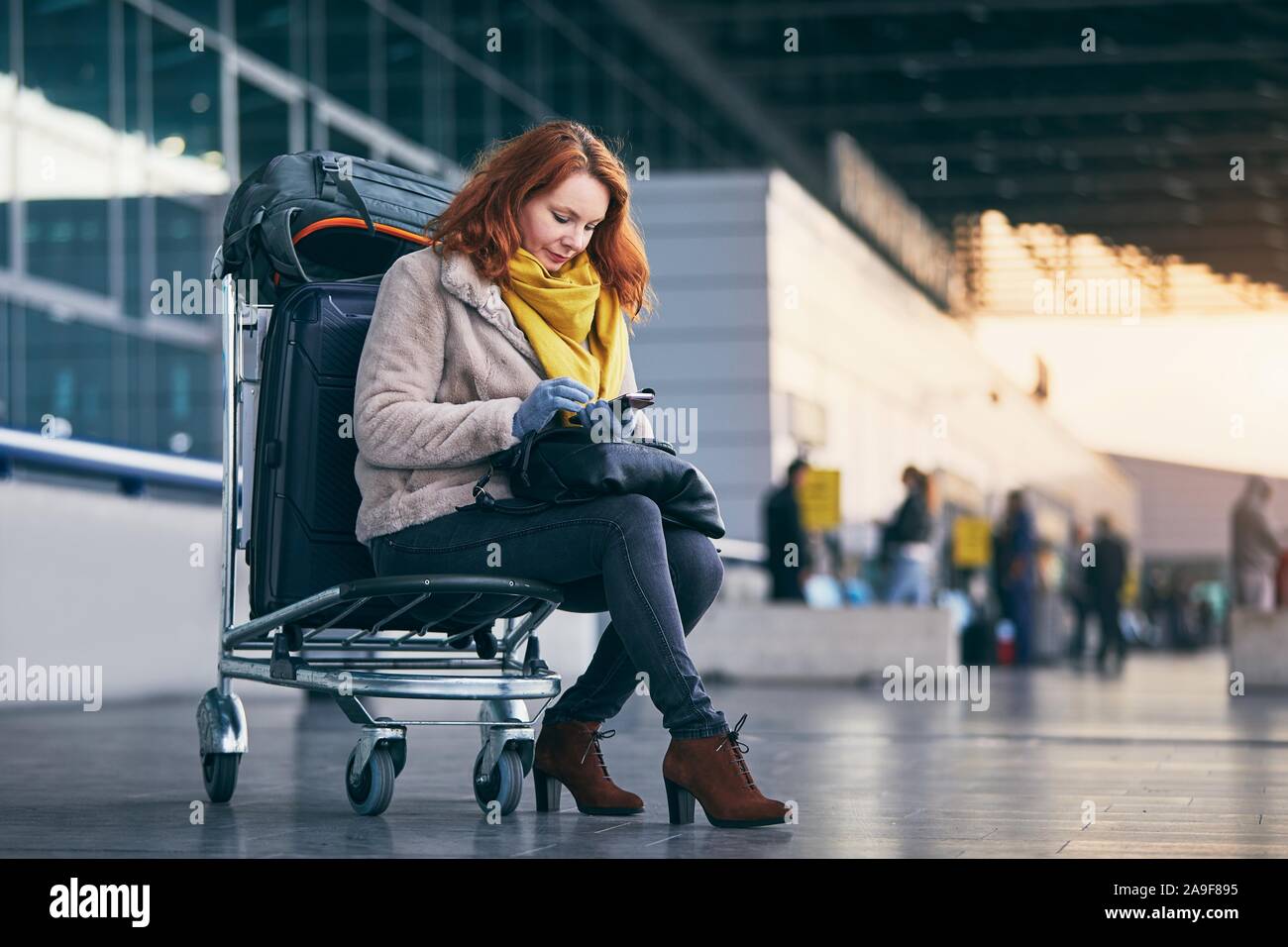 Junge Frau wartet auf Wagen mit Gepäck und mit smart phone gegen Flughafen Terminal Gebäude. Stockfoto