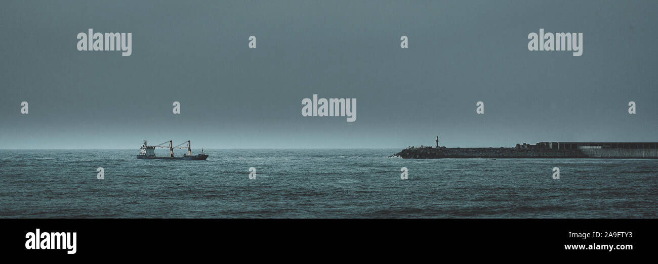 Frachtschiff in Port in der Nähe des Leuchtturm und Mole im äußeren Hafen. Bei Sonnenuntergang an einem dunklen Tag Stockfoto Frachtschiff in Port in der Nähe des Leuchtturm und Mole im äußeren Hafen. Bei Sonnenuntergang an einem dunklen Tag Stockfoto