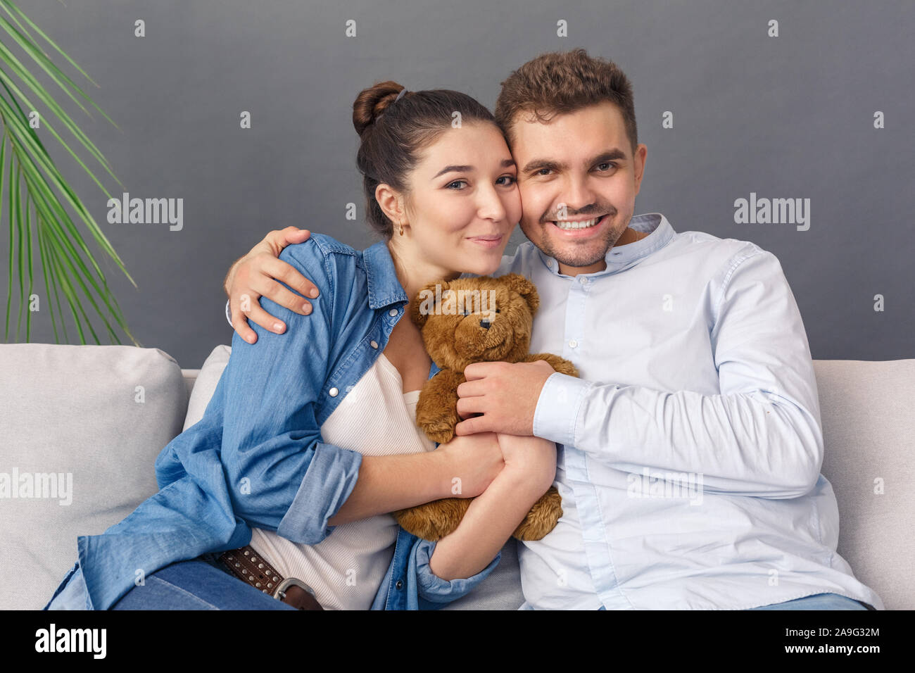 Relationnship Konzept. Junges Paar sitzt auf einem Sofa studio isoliert auf Grau mit Teddybär lächelnd Frohe Stockfoto