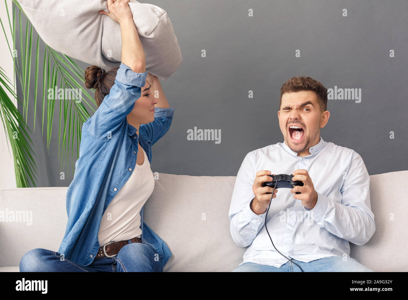Relationnship Konzept. Junges Paar Frau schlagen mit Kissen Mann spielt Spielekonsole schreien Sitzen auf einem Sofa studio isoliert auf Grau Stockfoto