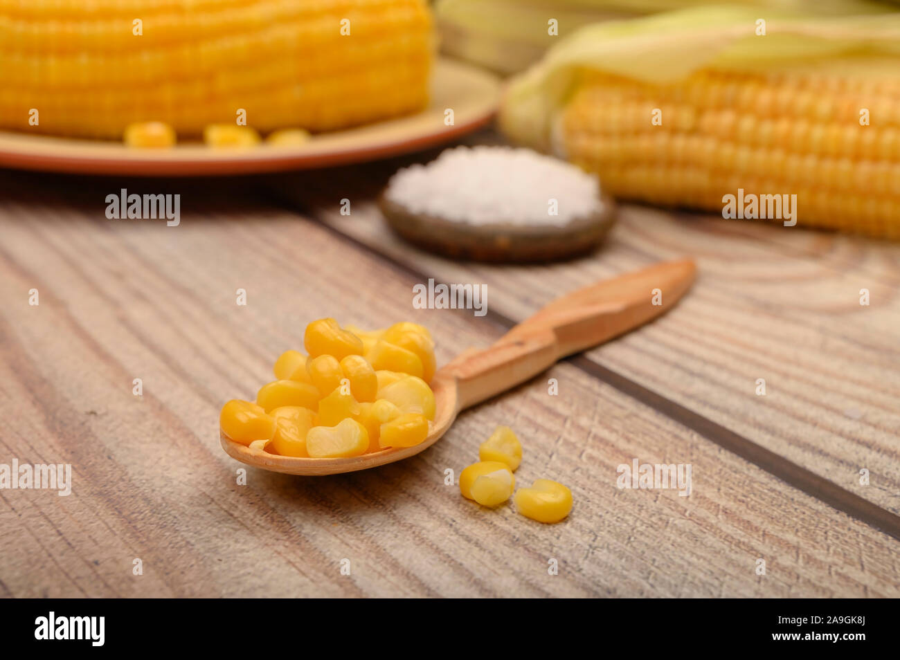 Körner von Mais auf einem Löffel aus Holz, auf einem Holztisch, im Hintergrund gekochten Mais, grobes Salz und unbehandelte Maiskolben. Fitness Ernährung. Gesund Stockfoto