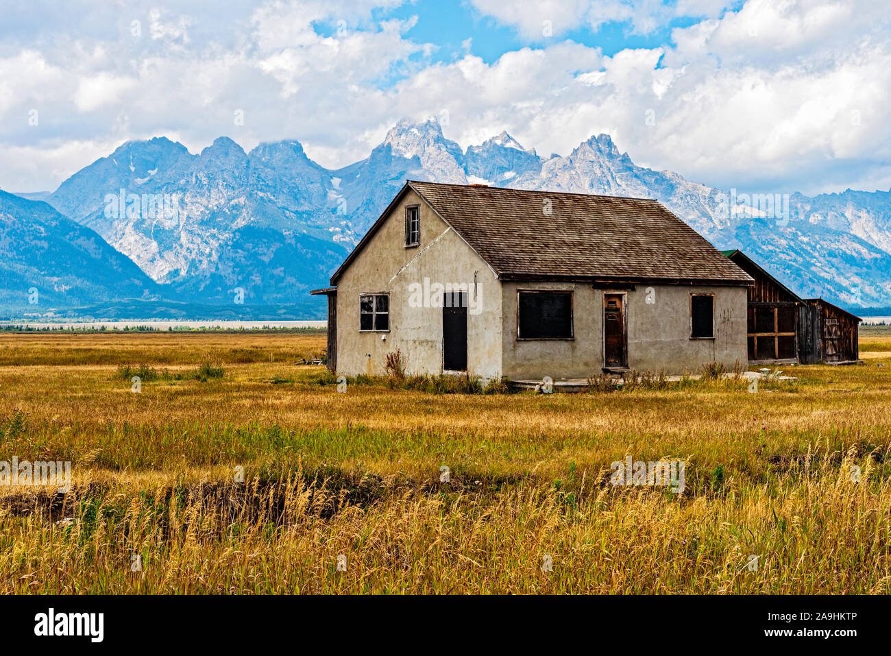Verlassenes Haus der Pionierfarm auf grasbewachsenem Feld mit hohen Rocky Mountains jenseits des blauen Himmels mit weißen, flauschigen Wolken. Stockfoto