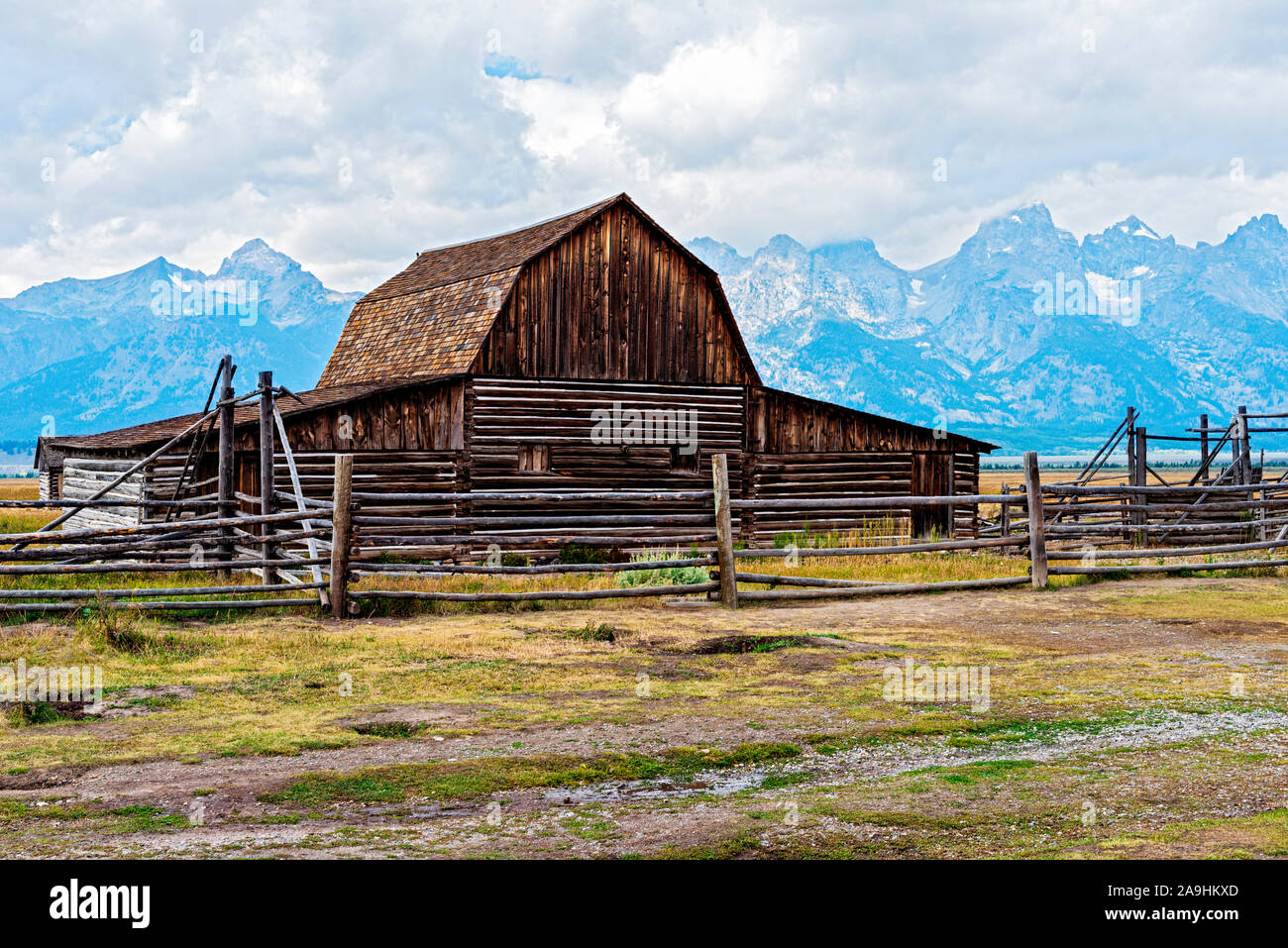 Alte Holzscheune mit eingezäunten Feldern und hohen Rocky Mountains jenseits des wolkenlosen Himmels. Stockfoto