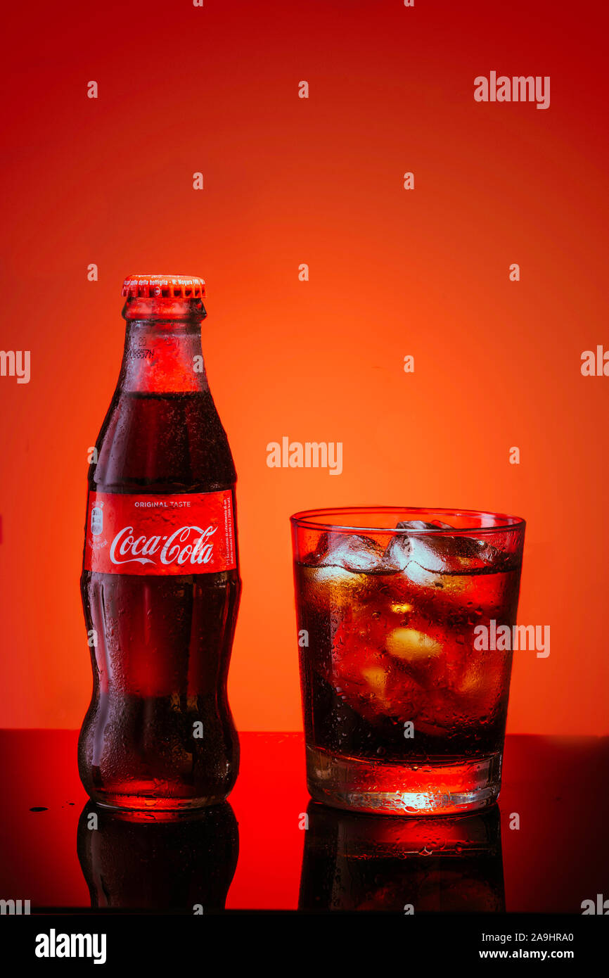 Italien, Mailand - 15. November 2019: Classic Flasche Coca-Cola und Glas frischen Coca Cola mit Eiswürfeln und Wassertropfen auf den Spiegel. Studio gedreht. Roter Hintergrund Stockfoto