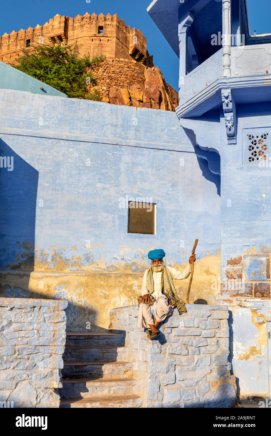 Älterer Mann in einem blauen Turban, der an einer Wand in der Blauen Stadt Jodhpur sitzt, mit dem Fort Mehrangarh im Hintergrund, Rajasthan, Indien Stockfoto