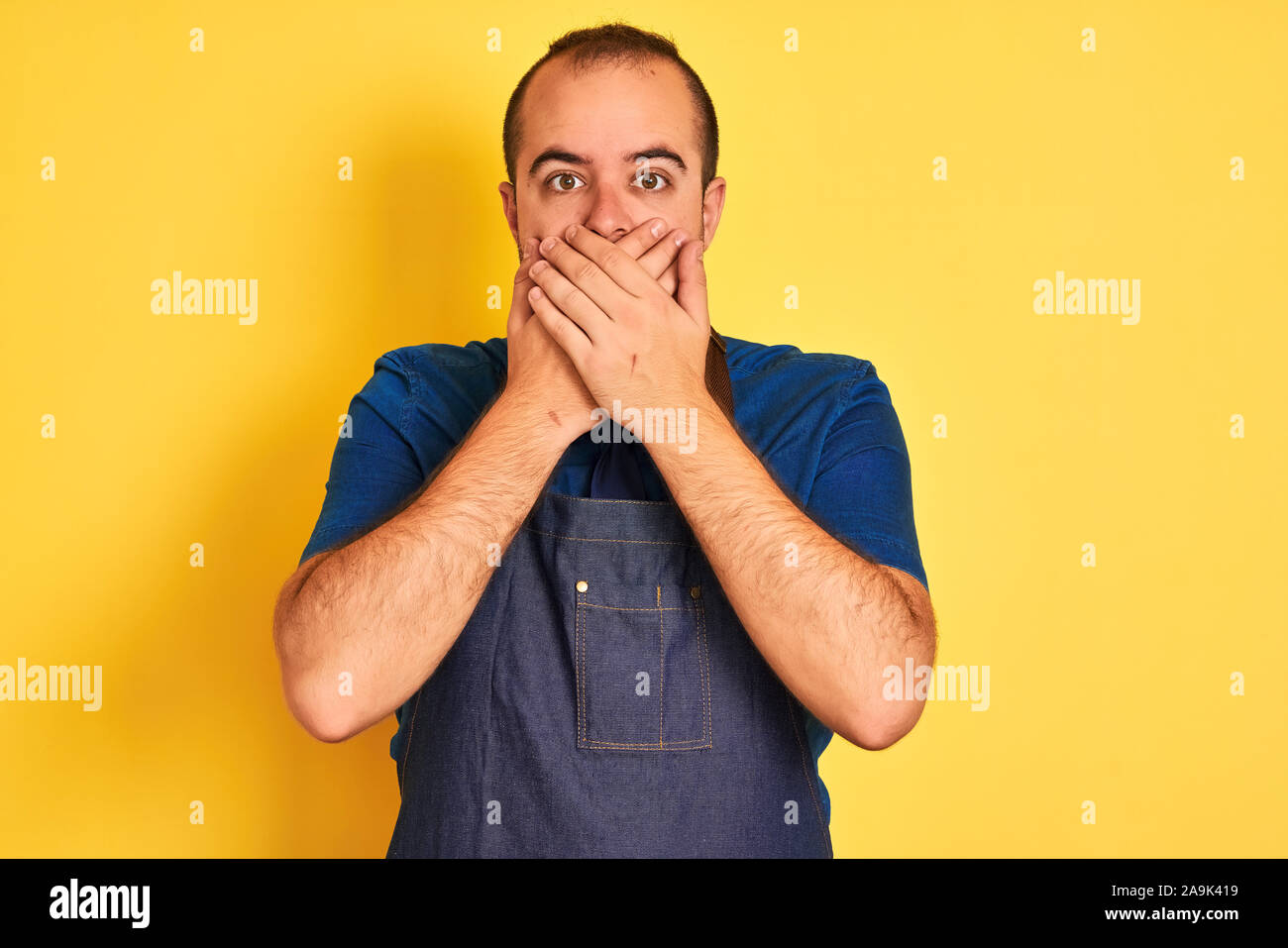Junge Ladenbesitzer Mann mit Schürze stehend über isolierte gelben Hintergrund schockiert über den Mund mit Händen für Fehler. Geheime Konzept. Stockfoto