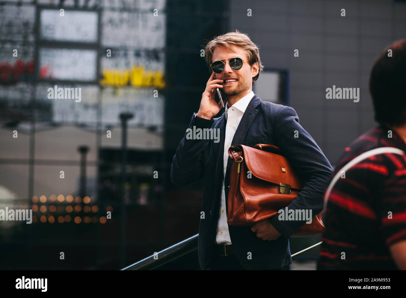Ein Mann mittleren Alters, der arbeitet in einem Büro ist zu Fuß entlang der Straße mit einer Aktentasche in der Hand und sprechen für eine Mobile und lächelnd Stockfoto