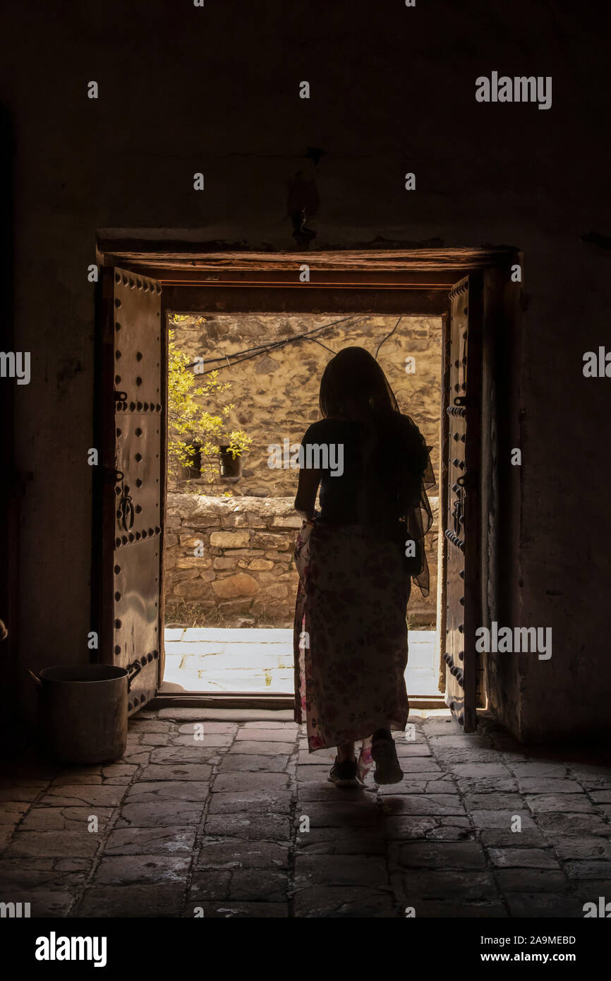 Beschattete Frau mit Kopftuch geht aus dunklen Gebäude in hellen Innenhof durch die antiken Türen Stockfoto