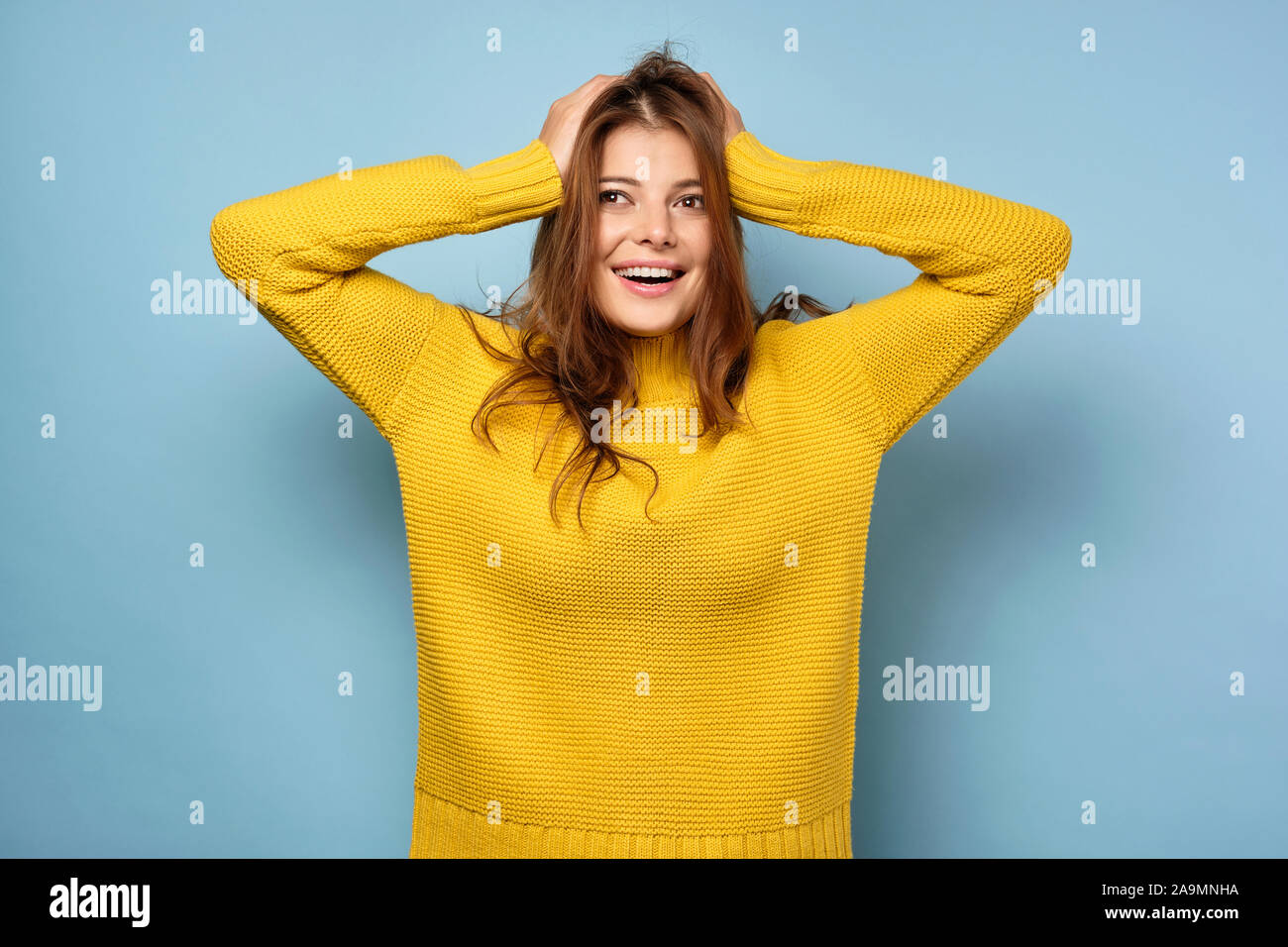 Die brünette steht auf einem blauen Hintergrund in einem gelben Pullover und schaut auf die Seite, ihr Haar ruffling mit ihren Händen. Stockfoto