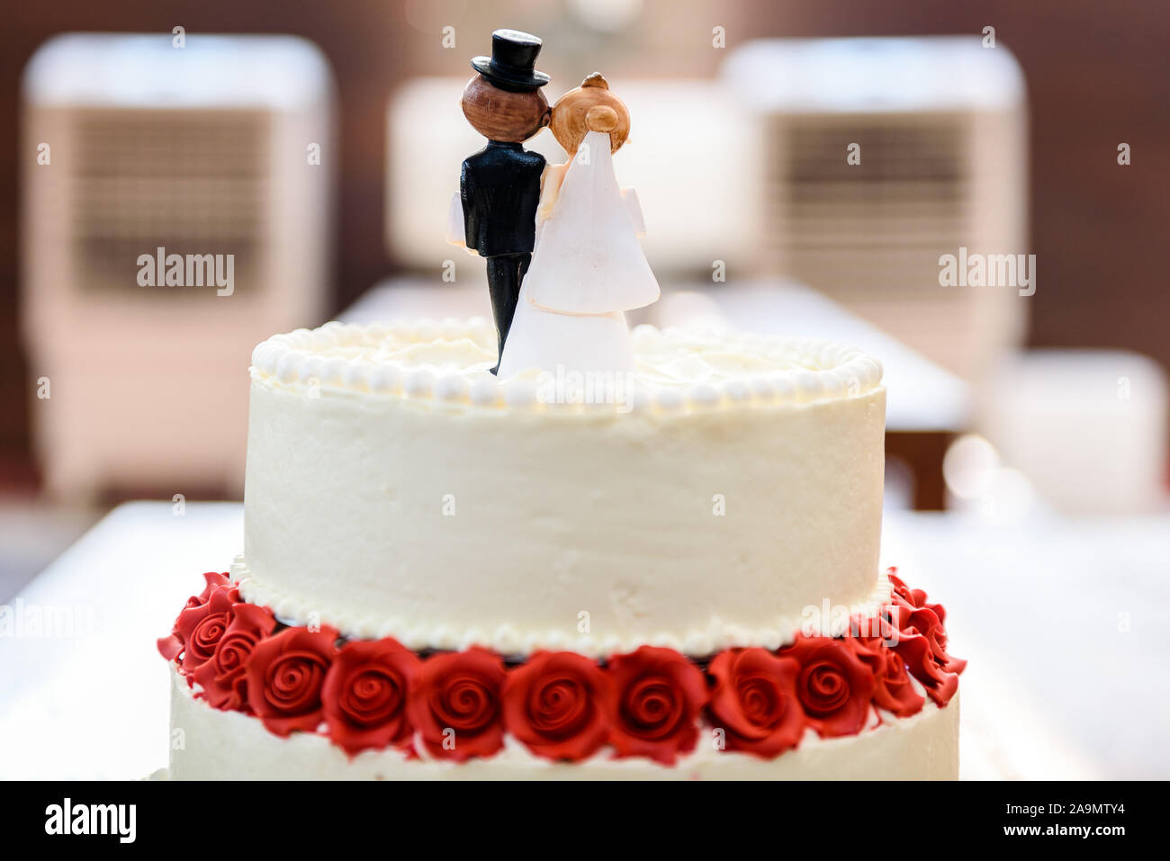 Weisse Hochzeitstorte mit Braut und Bräutigam Figuren an der Spitze. Schöne weisse Hochzeitstorte mit roten Rosen in der Tabelle mit den kleinen Mann und Frau Marionetten Stockfoto