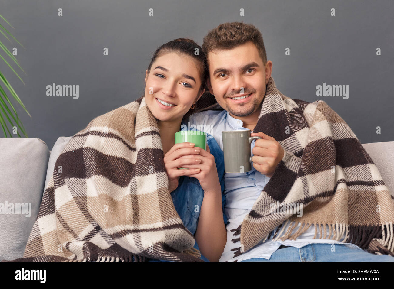 Relationnship Konzept. Junges Paar sitzt auf einem Sofa umarmen mit Decke Studio auf Grau trinken heißen Tee glücklich lächelnd in die Kamera isoliert abgedeckt Stockfoto