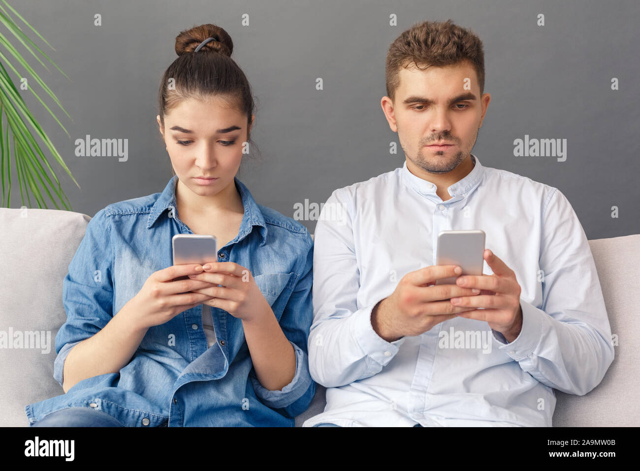 Moderne Relationnship Konzept. Junges Paar sitzt auf einem Sofa Studio auf Grau surfen Smartphone isoliert konzentriert Stockfoto