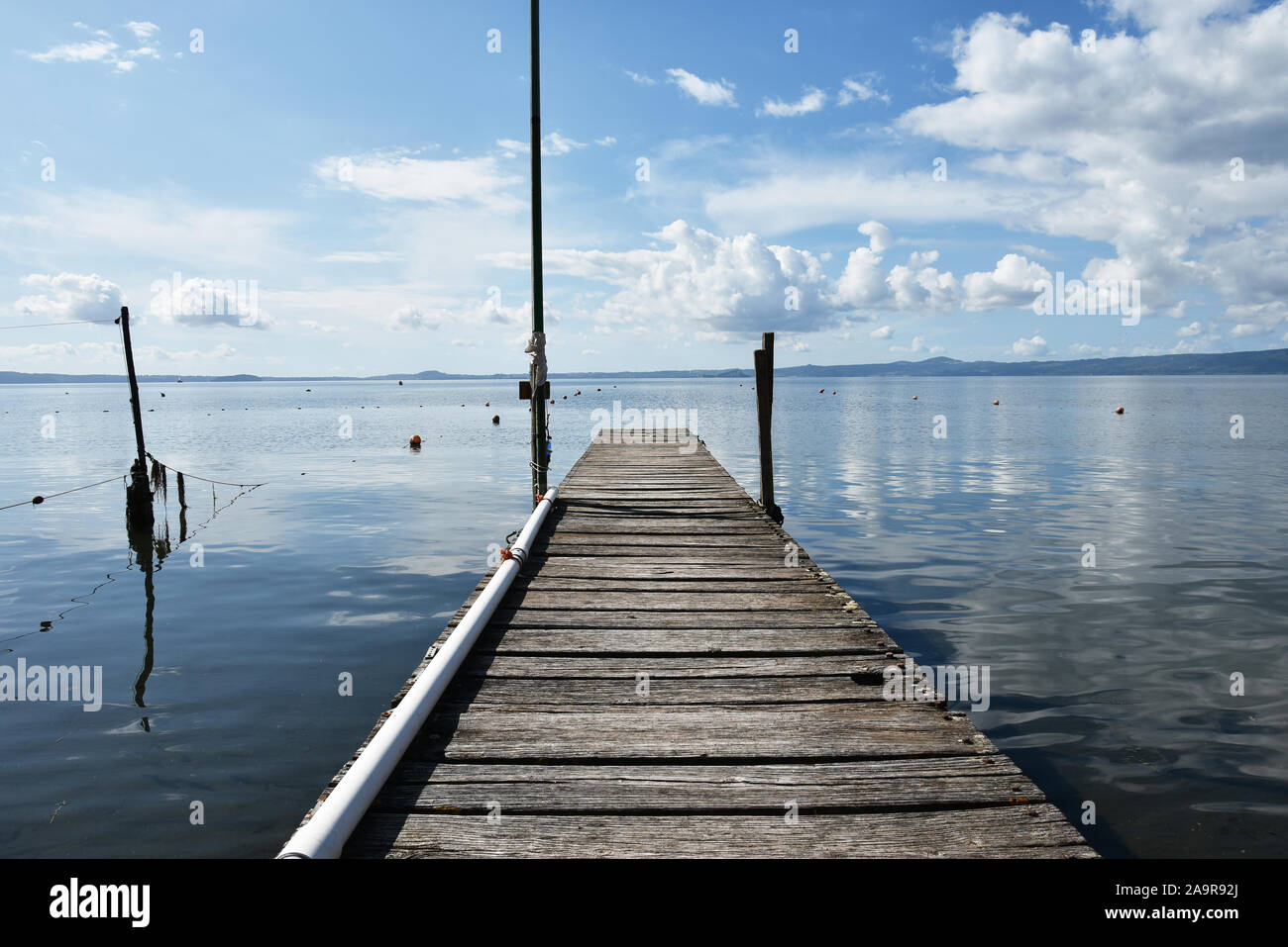 Hölzerne Anlegesteg für Boote vor blauem Himmel mit einigen Wolken. Bojen und Fischernetze. Hügeln im Hintergrund. Bolsena See, Italien. Stockfoto