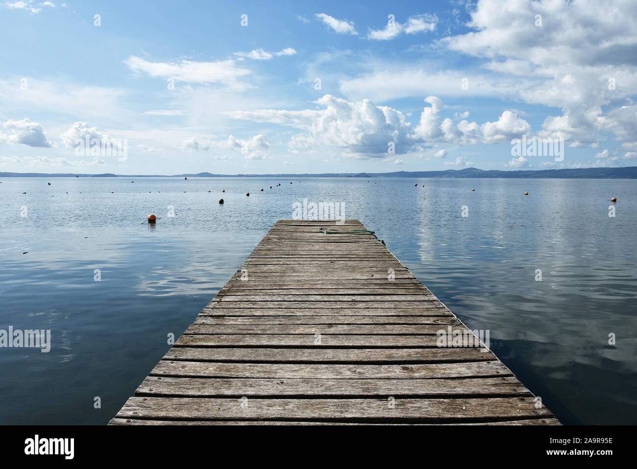 Hölzerne Anlegesteg für Boote vor blauem Himmel mit einigen Wolken. Bojen. Hügeln im Hintergrund. Bolsena See, Italien. Stockfoto