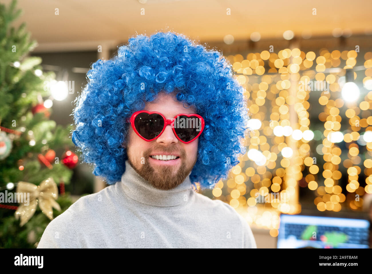 Fröhlicher junger Geschäftsmann, der in blauen Lockenperücke und heartshaped Sonnenbrille Stockfoto