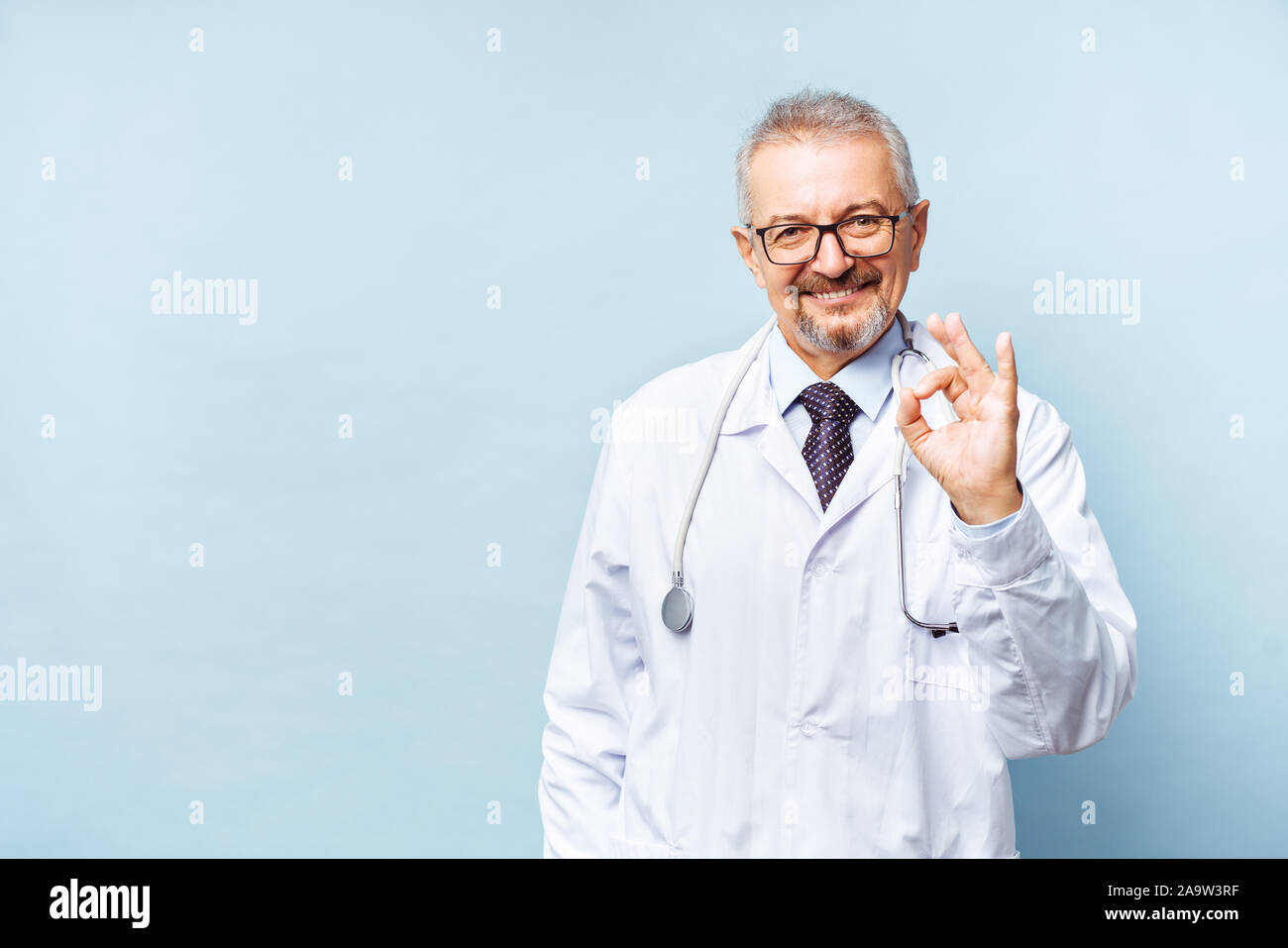 Fröhliche reife Arzt posing und lächelnd an Kamera, Gesundheitswesen und Medizin. Auf blauem Hintergrund isolieren. Arzt zeigt ein ok Stockfoto