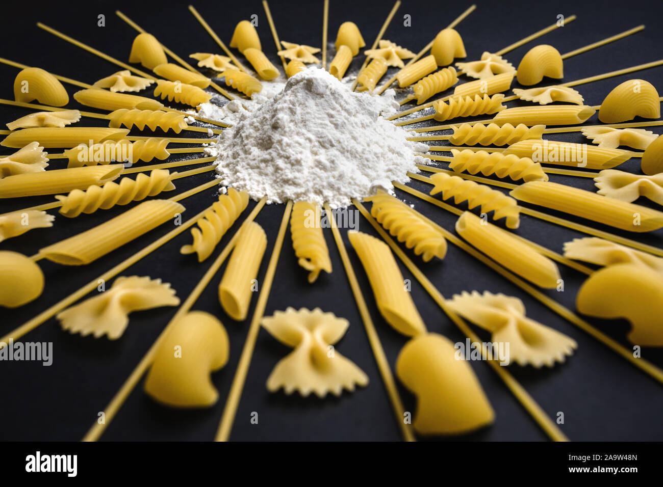 Pasta angeordnet um Stapel von Mehl und Weizen. Geometrische Anordnung unterschiedlicher Art der Nudeln auf schwarzem Hintergrund. Stockfoto